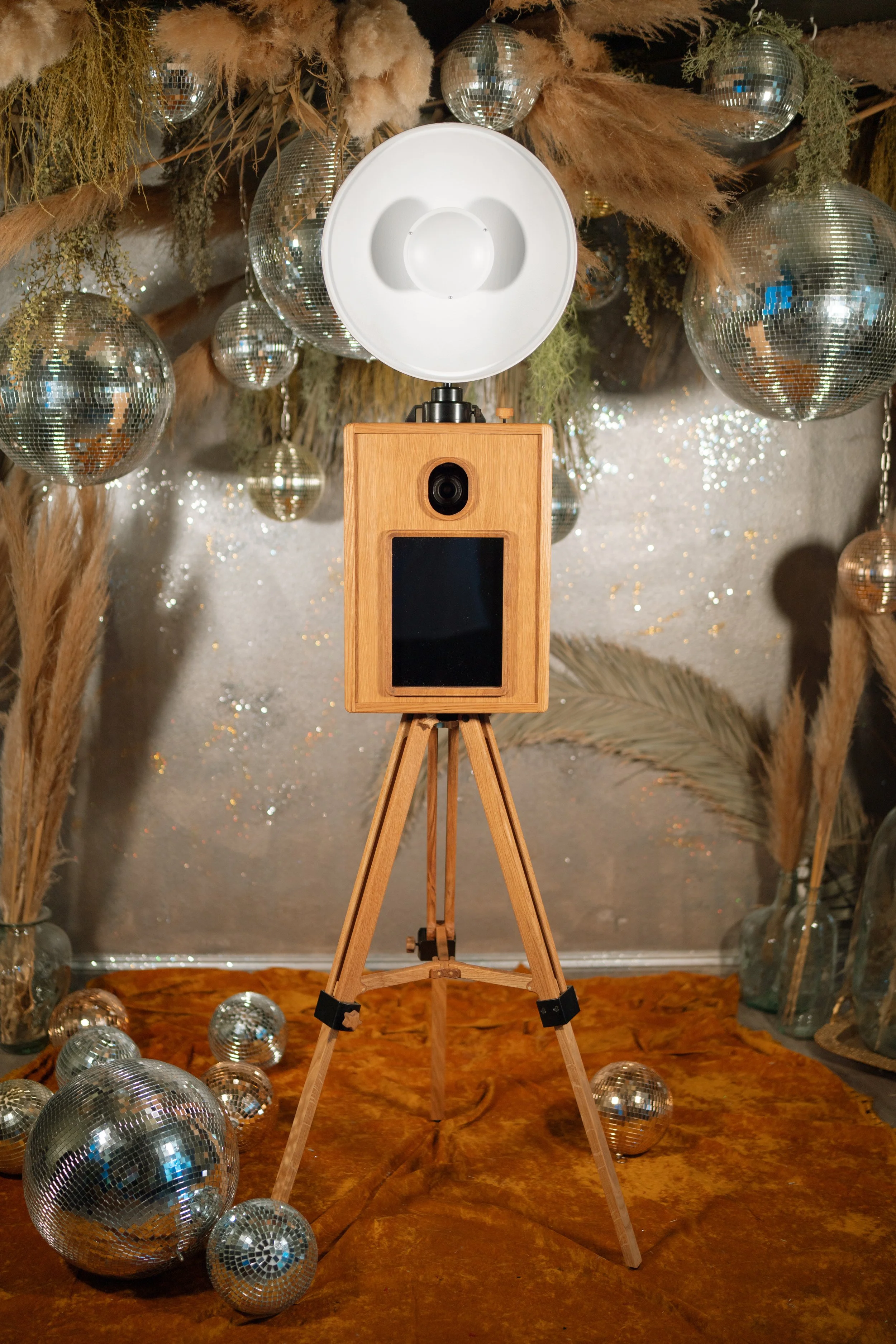A wooden photo booth on a tripod stand surrounded by disco balls and dried plants, with a silver glittery background.