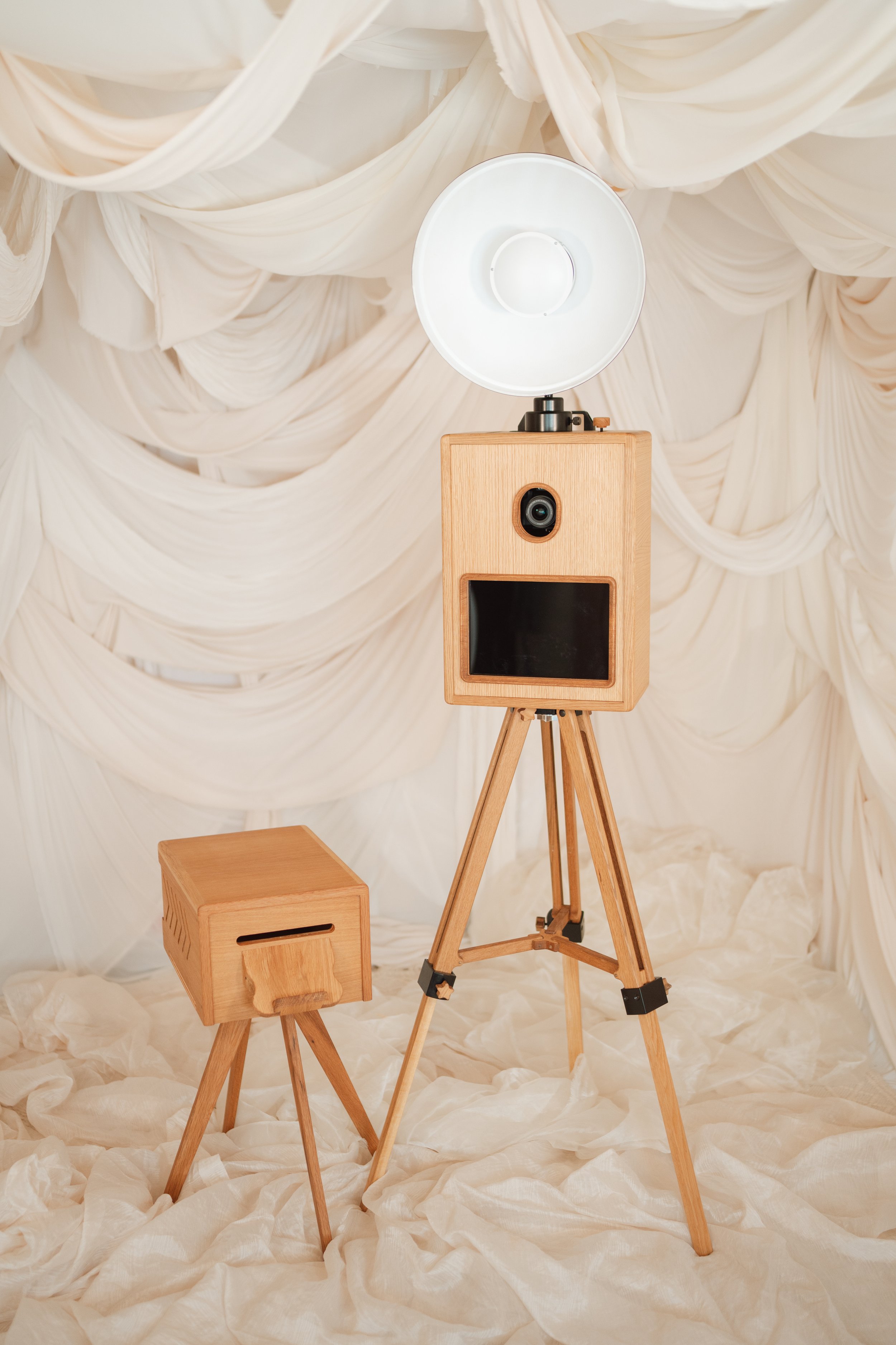 A wooden photo booth with a large flat screen monitor, a camera above it, and a large lamp on top, standing on a tripod. A smaller wooden box on four legs is beside it, all set against a cream-colored, draped fabric backdrop.