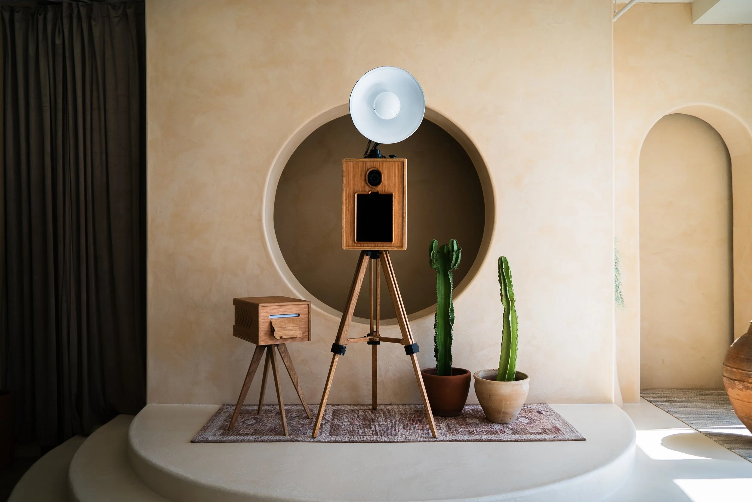 Interior space with beige textured walls, a circular cutout in the wall, a vintage wooden floor lamp with a large reflector lamp, two potted cacti, a small wooden side table, a beige rug, and a dark curtain on the left.