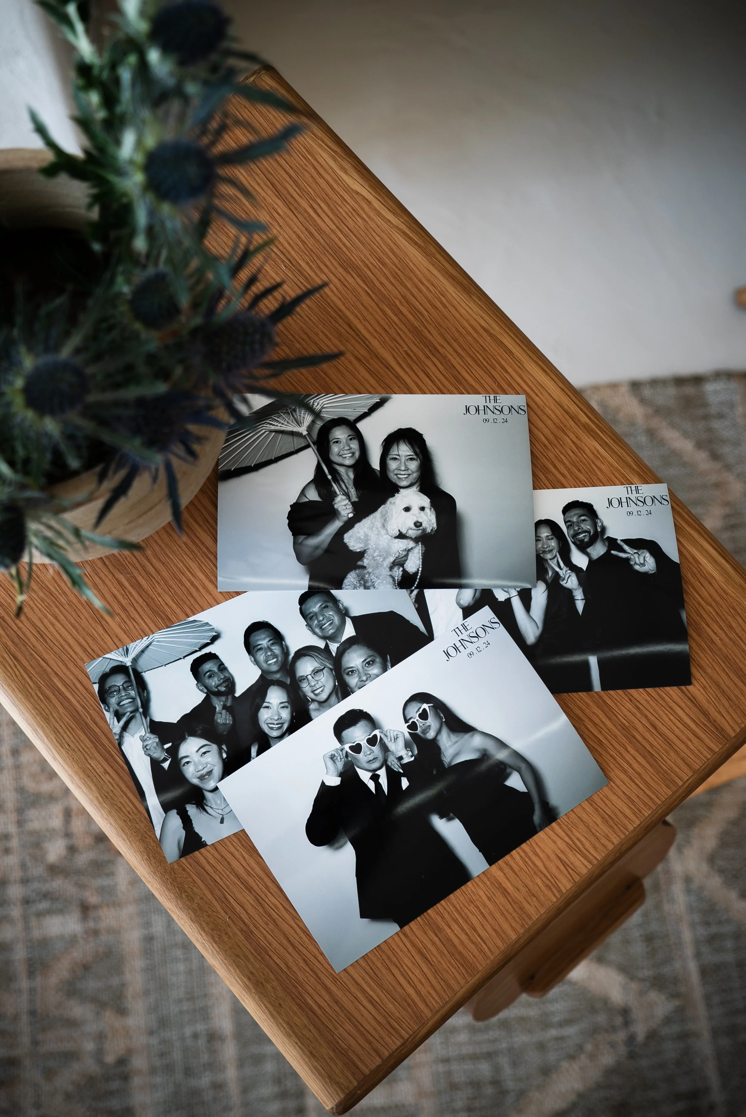 Black and white photo booth pictures of a group of people at a celebration, some posing with props like sunglasses and umbrellas, on a wooden surface with a vase of dried flowers partially visible at the top left corner.