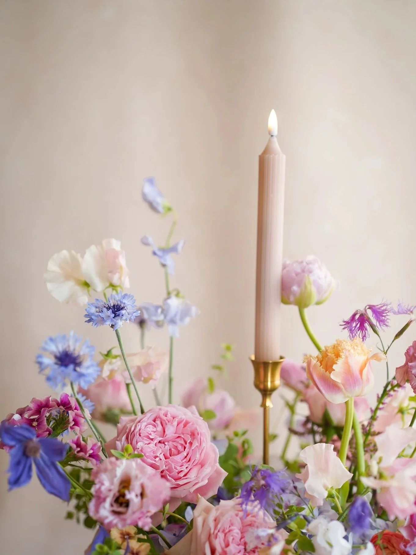 A pink candle burning on a brass holder surrounded by pastel-colored flowers including roses and delphiniums, with a neutral background.