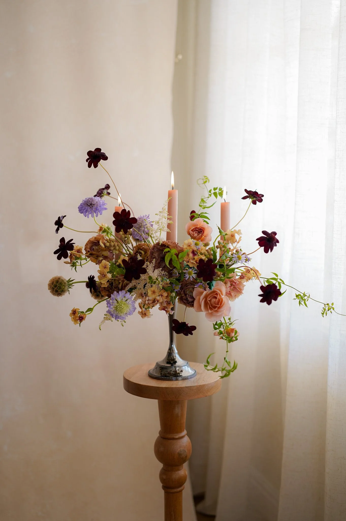 A floral arrangement with pink roses, purple and dark red flowers, and greenery, placed in a silver vase with two lit pink candles, on a wooden stand near a sheer curtain.