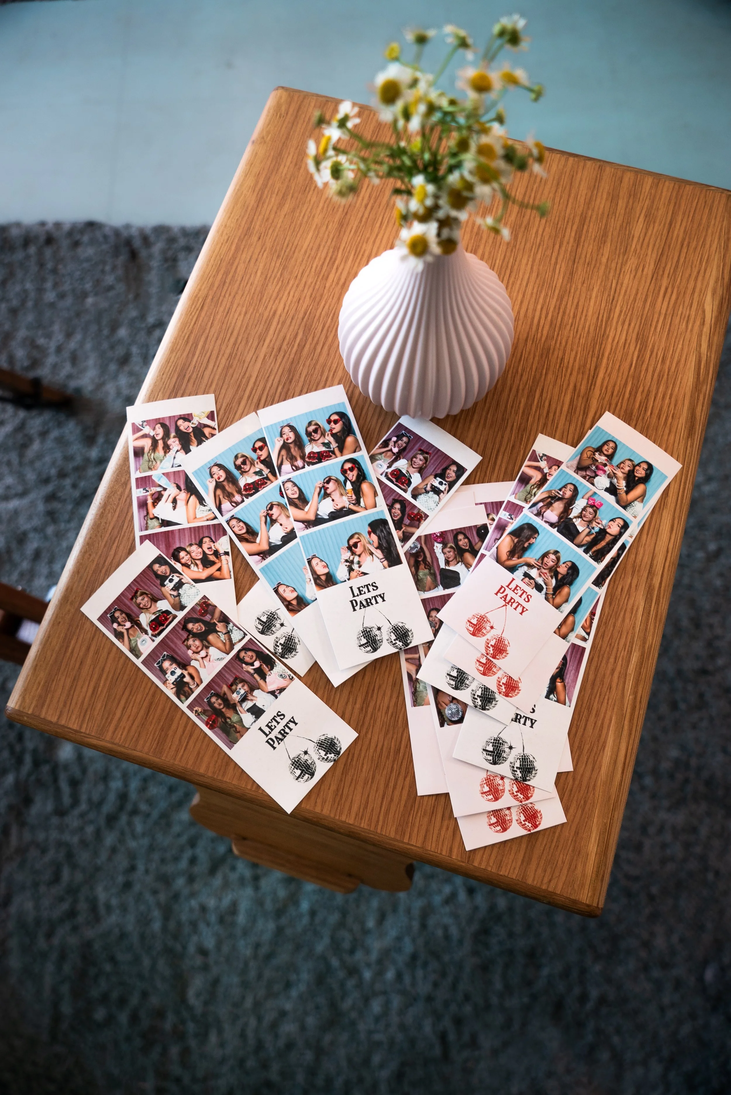 Photo booth photo strip with four pictures of a group of young women posing and having fun. The photo strip has a caption that says 'Let's Party' and a graphic of disco balls. The photo strip is on a wooden table next to a white vase with a textured surface, holding a bouquet of small white and yellow flowers.