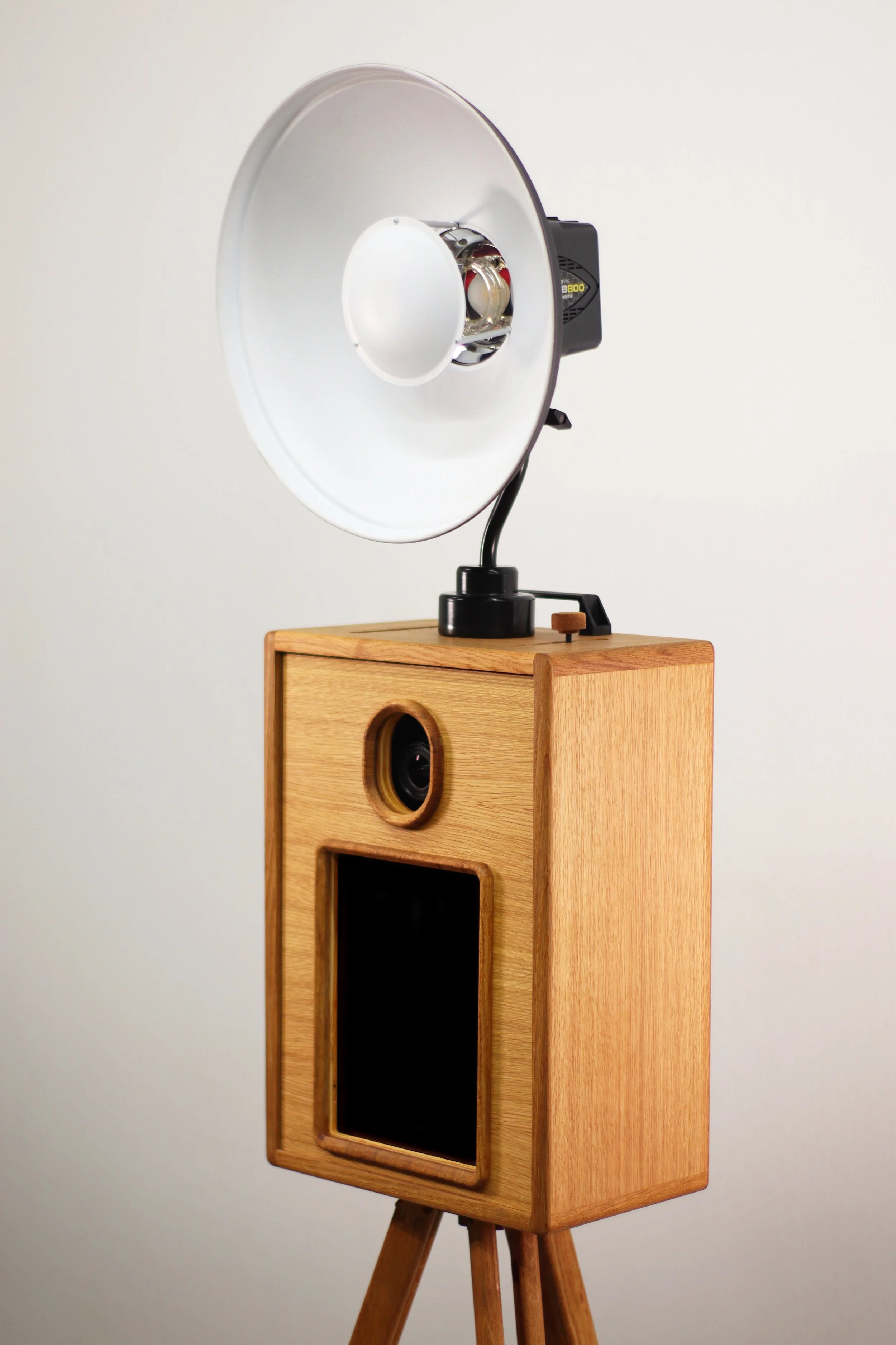 A vintage photo booth with a wooden exterior, a camera lens, and a black screen, topped with a large studio light on a stand, against a plain white background.