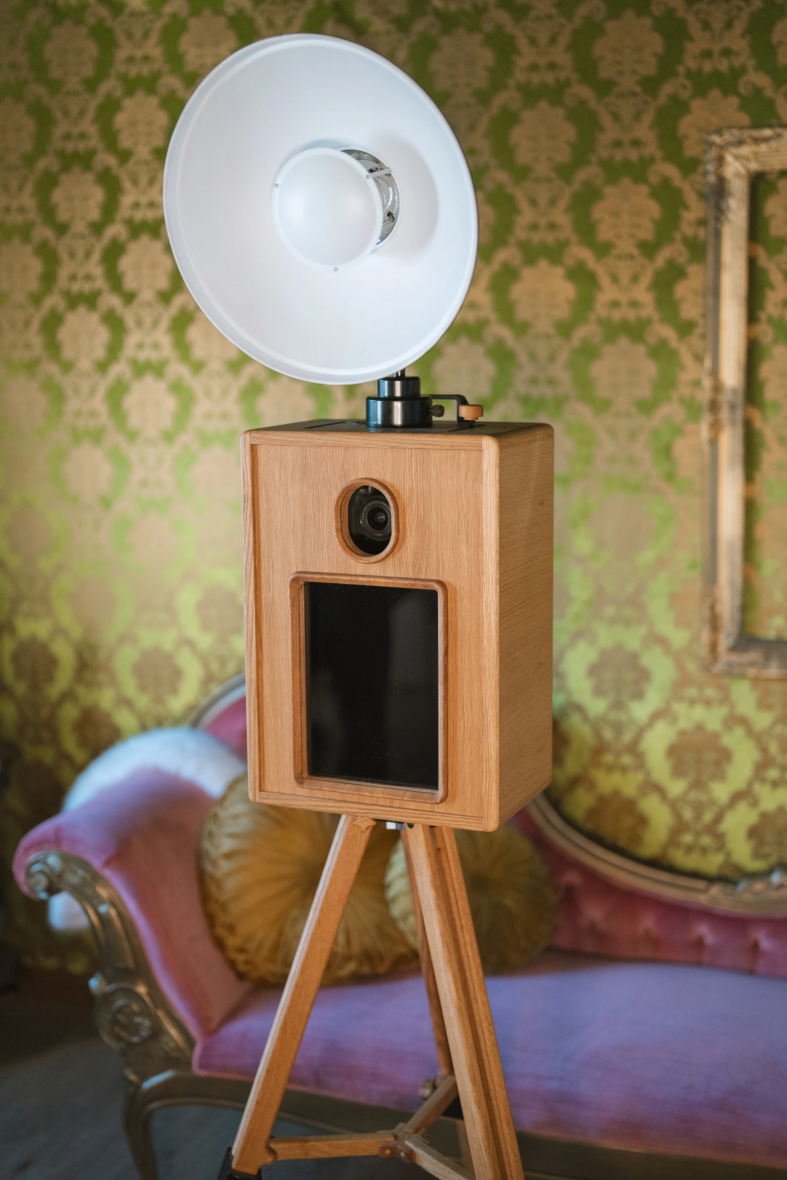 A vintage wooden camera with a large white flash on a tripod, positioned in front of a pink Victorian-style sofa with floral pillows, and patterned green and beige wallpaper.
