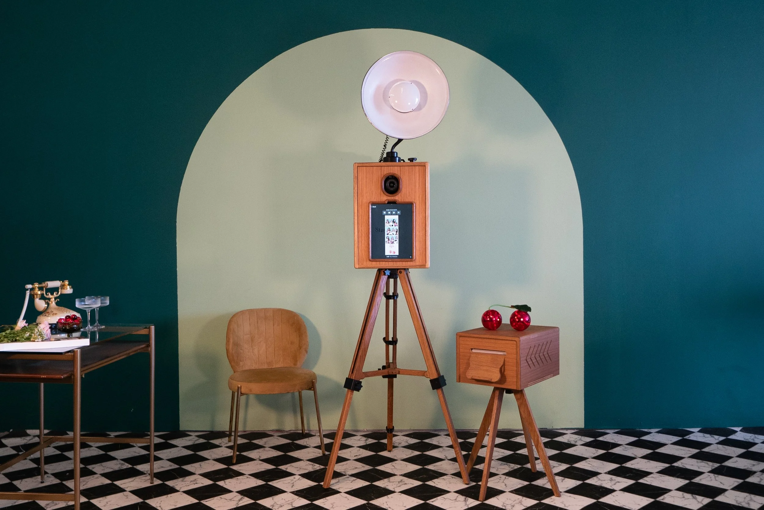 A room with a teal and light green wall, checkered black and white floor, and vintage furniture. There is a wooden chair, a small wooden side table with two red Christmas ornaments, a tall wooden tripod with a camera or device, and a vintage-style phone on a table. A large white circular light or reflector is mounted above the tripod.