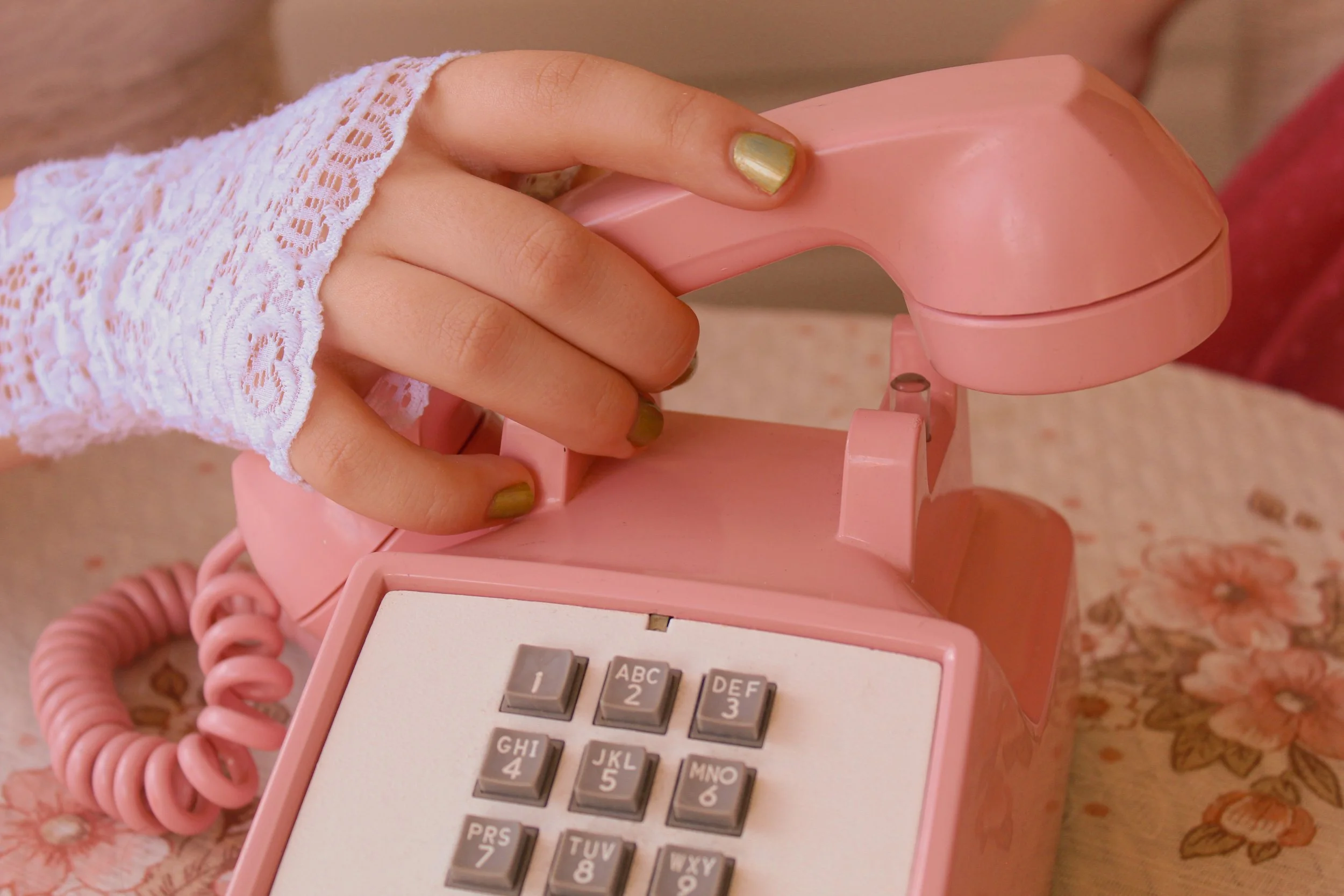 A person with green nail polish and lace sleeve holding a pink rotary phone, with the phone's handset lifted.