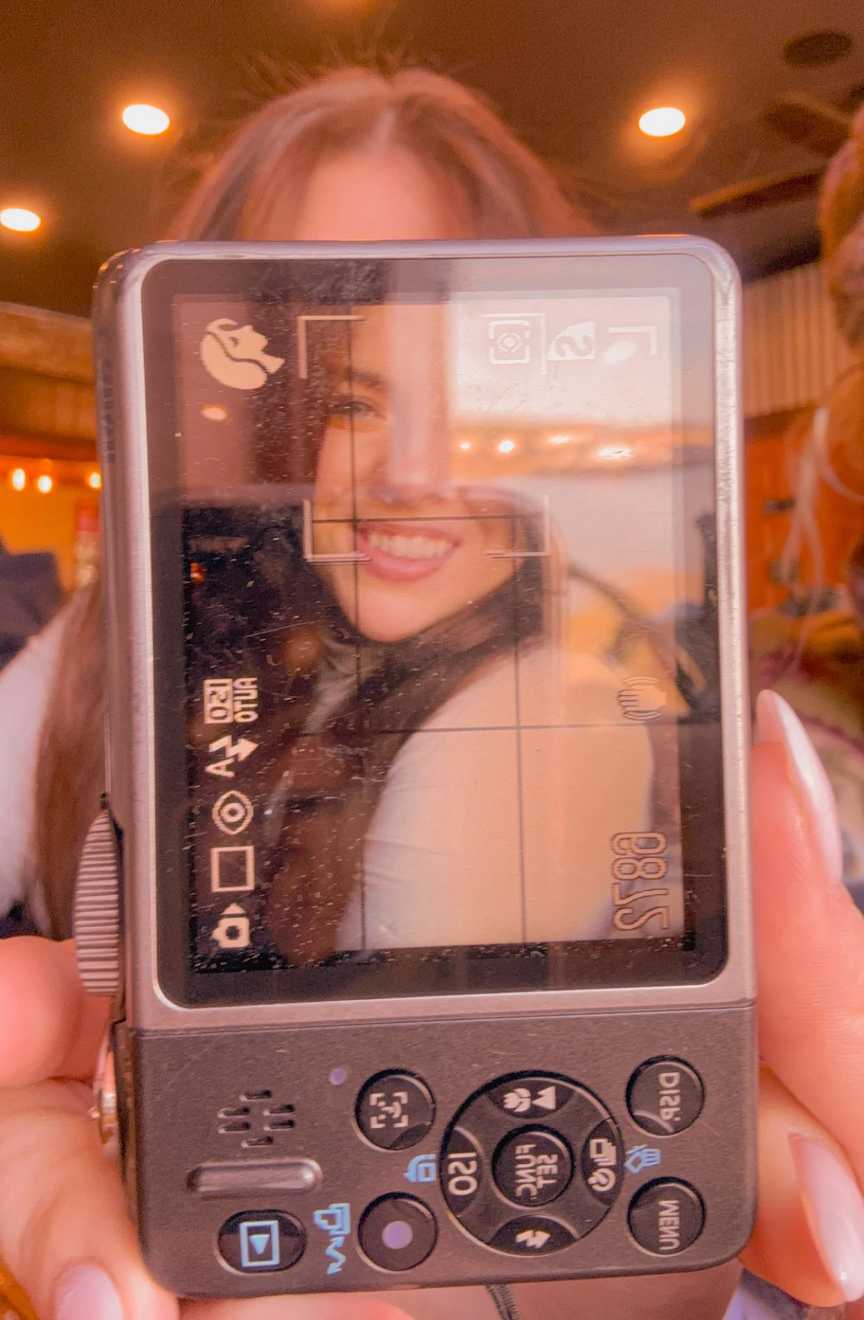 A woman taking a selfie with a digital camera, which shows her smiling face on the screen. The background appears to be a warmly lit indoor setting, possibly a restaurant or cafe.