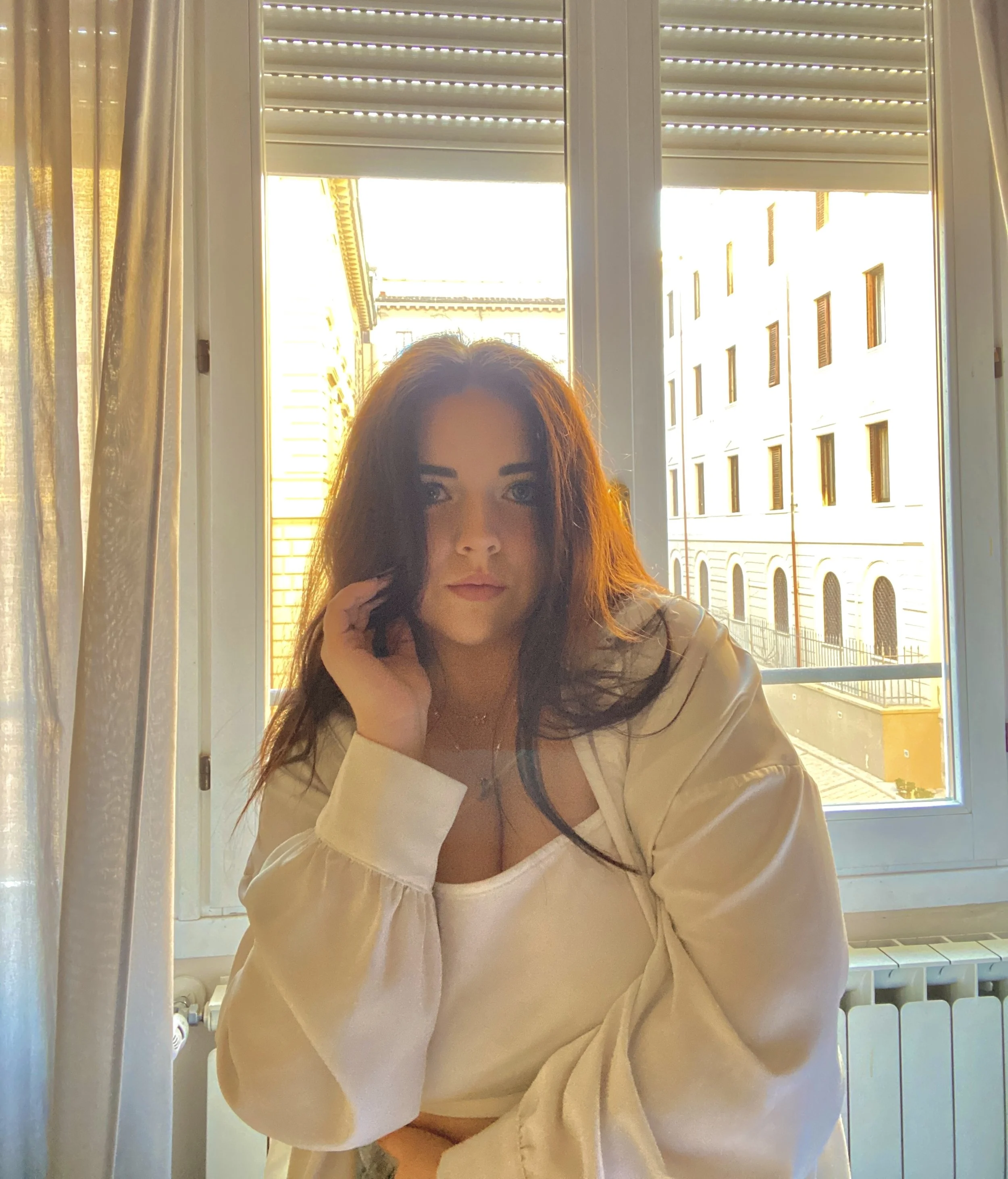 A woman with long dark hair sits in front of a window with partially open blinds, natural sunlight illuminating her face. She is wearing a cream-colored top and looking at the camera, with a city building visible outside the window.