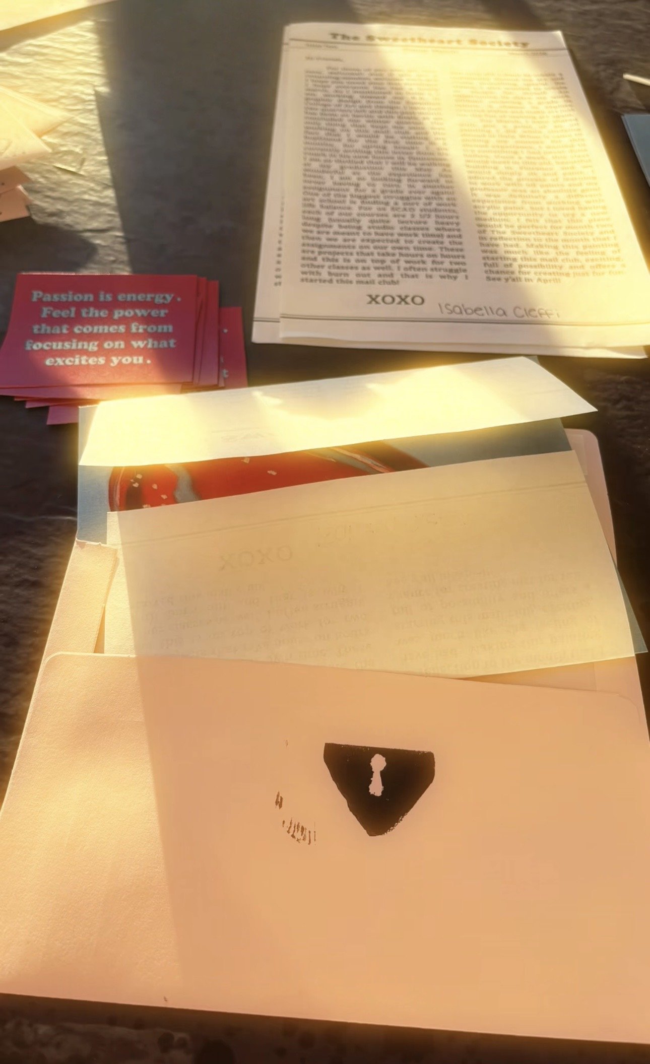 A table with various papers, including a printed program or article, some pink cards with a motivational quote, and a white envelope or folder with a black triangular symbol and a small punched hole. The scene is illuminated by warm sunlight.