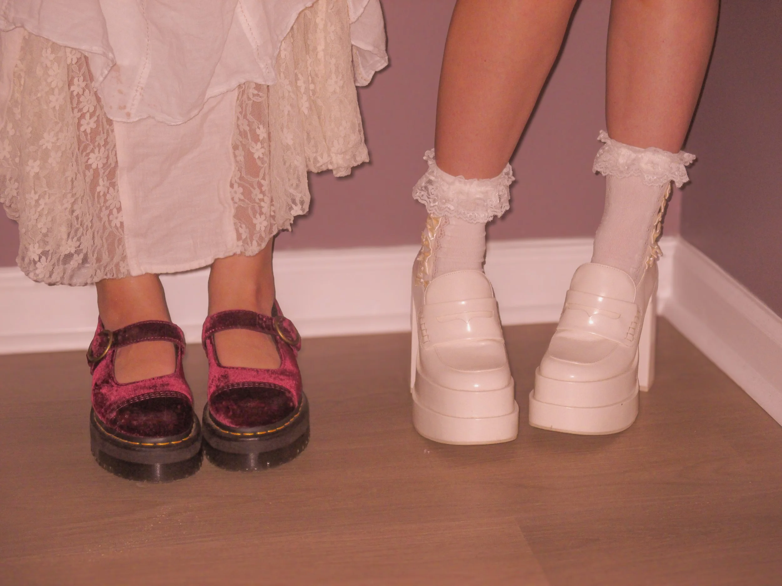 Close-up of two doll legs and shoes, both dolls dressed in lacy dresses. The doll on the left wears chunky pink velvet Mary Jane shoes, while the doll on the right wears shiny white platform shoes with lace-trimmed socks.