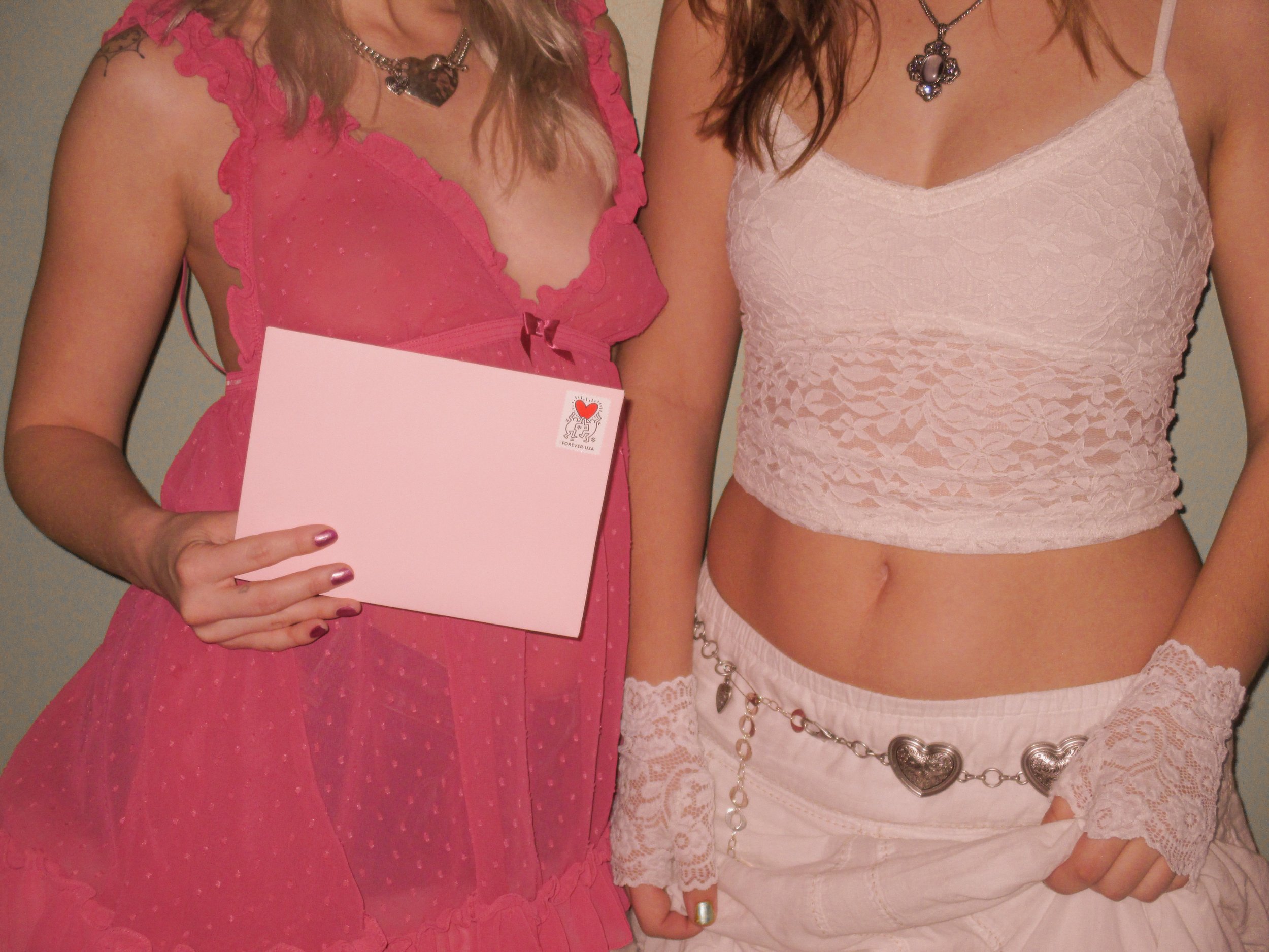 Two women in pink and white clothing with necklaces, one holding a pink card with a small stamp on it.