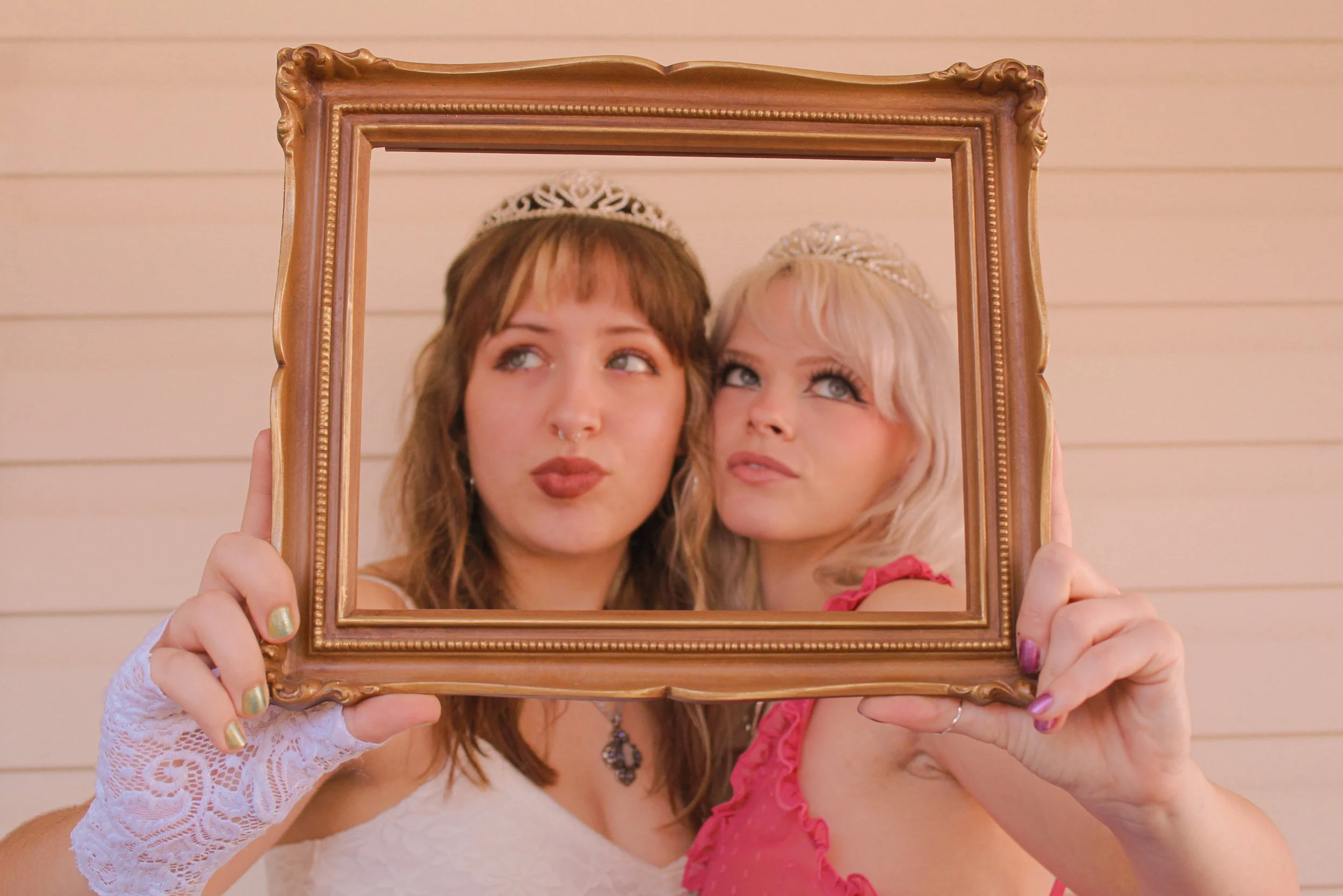 Two women wearing tiaras and makeup hold a decorative picture frame, framing their faces, while standing against a beige wall.