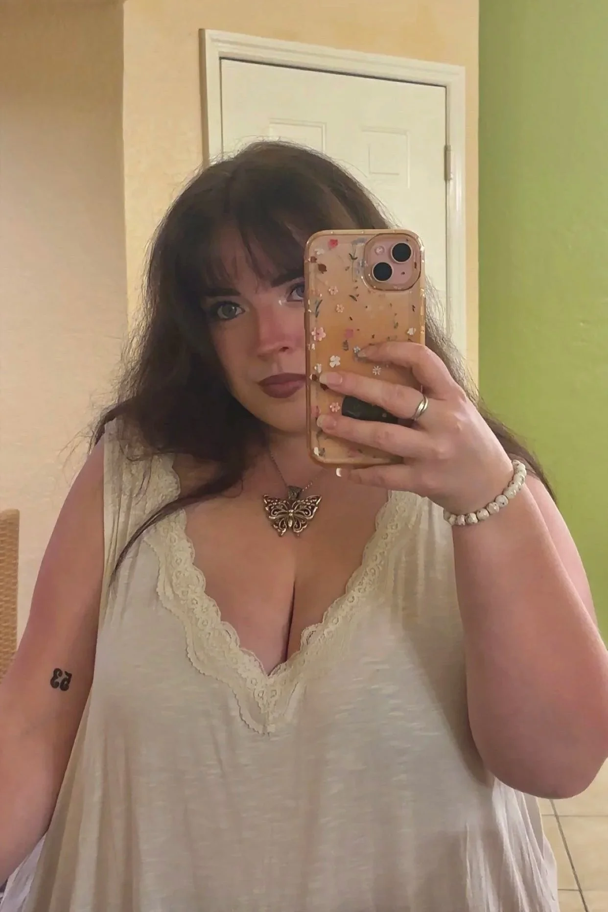 A woman taking a mirror selfie with a floral phone case in a room with beige and green walls. She has dark wavy hair, dark lipstick, a butterfly necklace, a bracelet, and a small tattoo on her upper left arm.