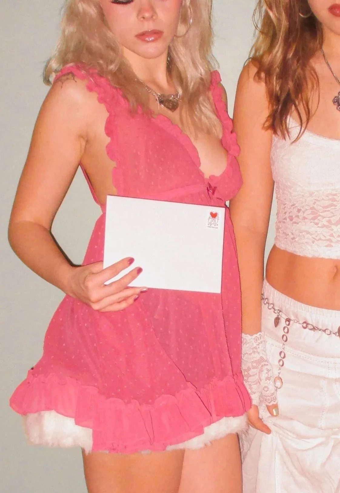 Young woman in a pink dress holding a white envelope with a small red heart in the corner, standing next to another person wearing a white crop top and chain accessories.