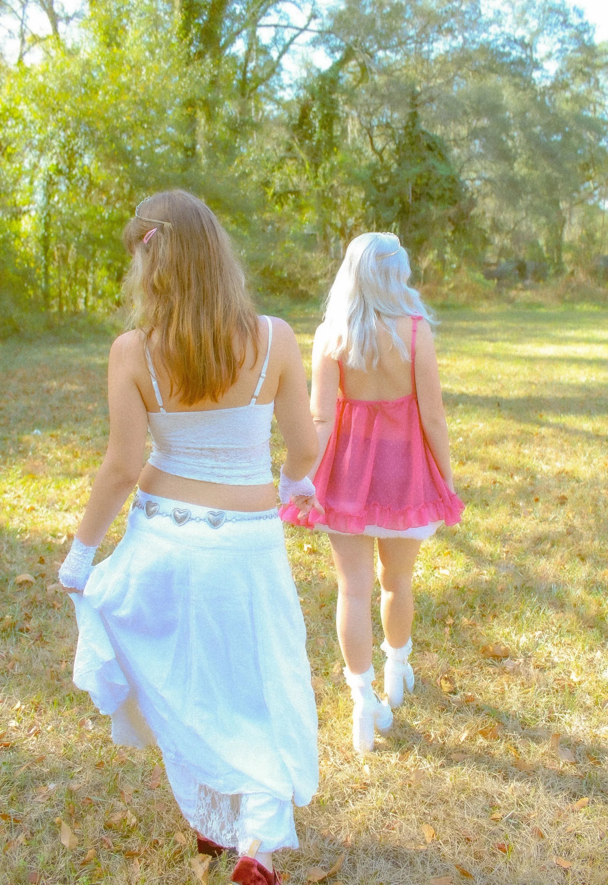 Two young women walking in a sunny park, holding hands, with trees and grass around them.