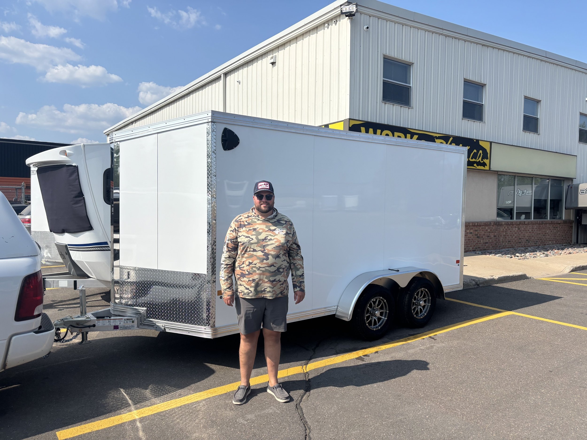A man stands in front of a white enclosed trailer attached to a vehicle in a parking lot, with a building and a clear blue sky in the background.