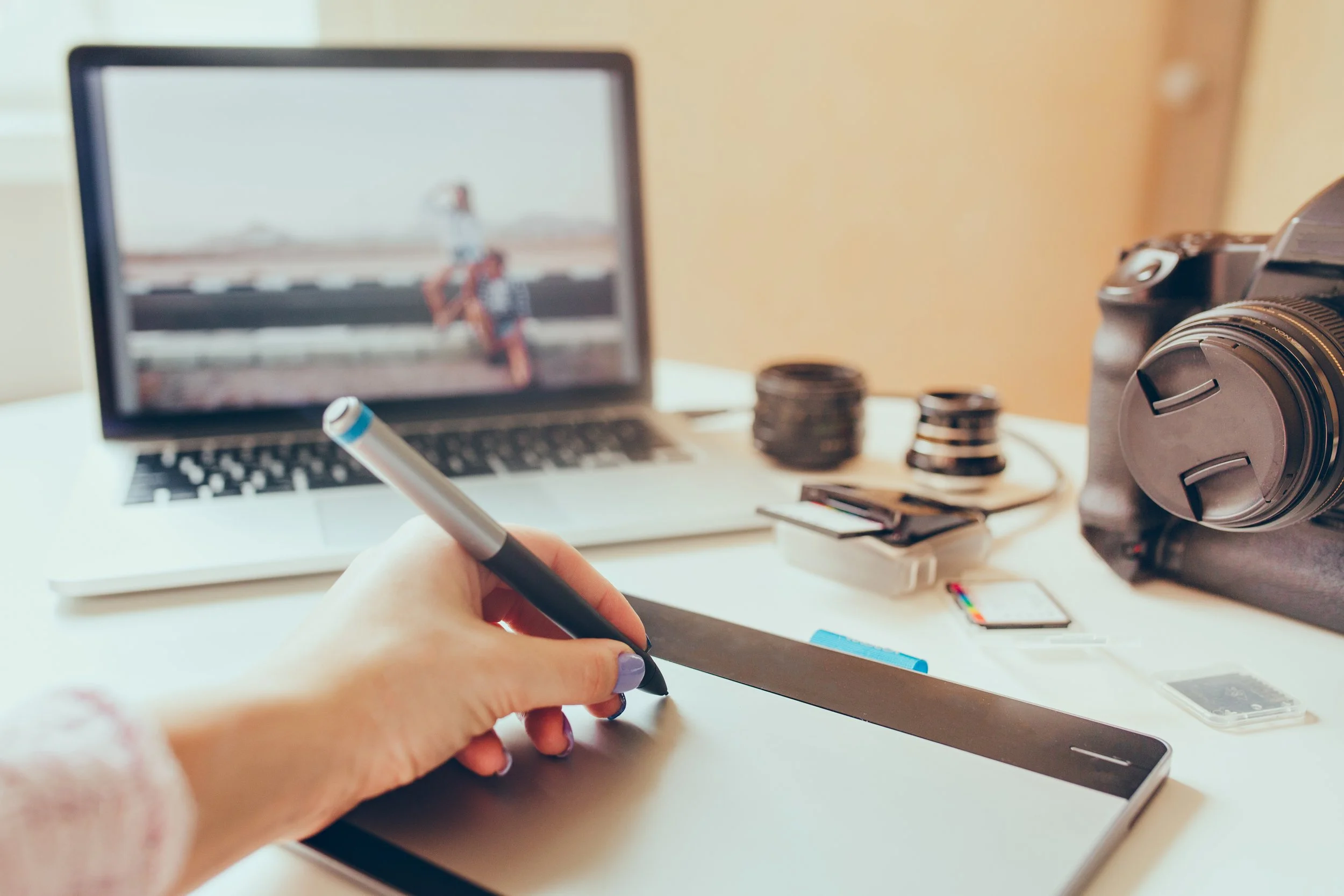 A person using a stylus on a graphics tablet, with a camera, a laptop, and camera accessories on a desk, and a blurred image of two people sitting on a bench on a laptop screen.