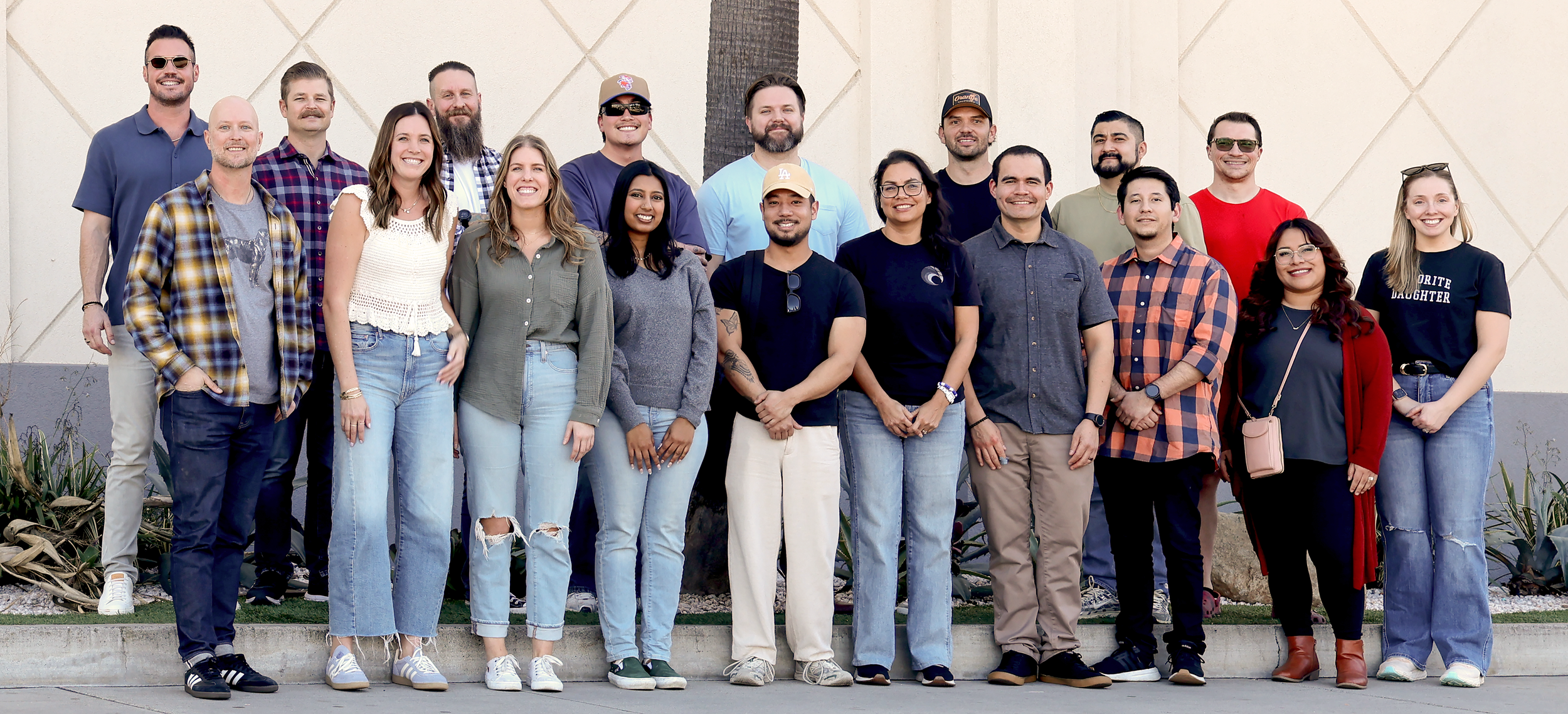 A group of 18 diverse people standing together outdoors, smiling, in front of a beige wall with a palm tree.