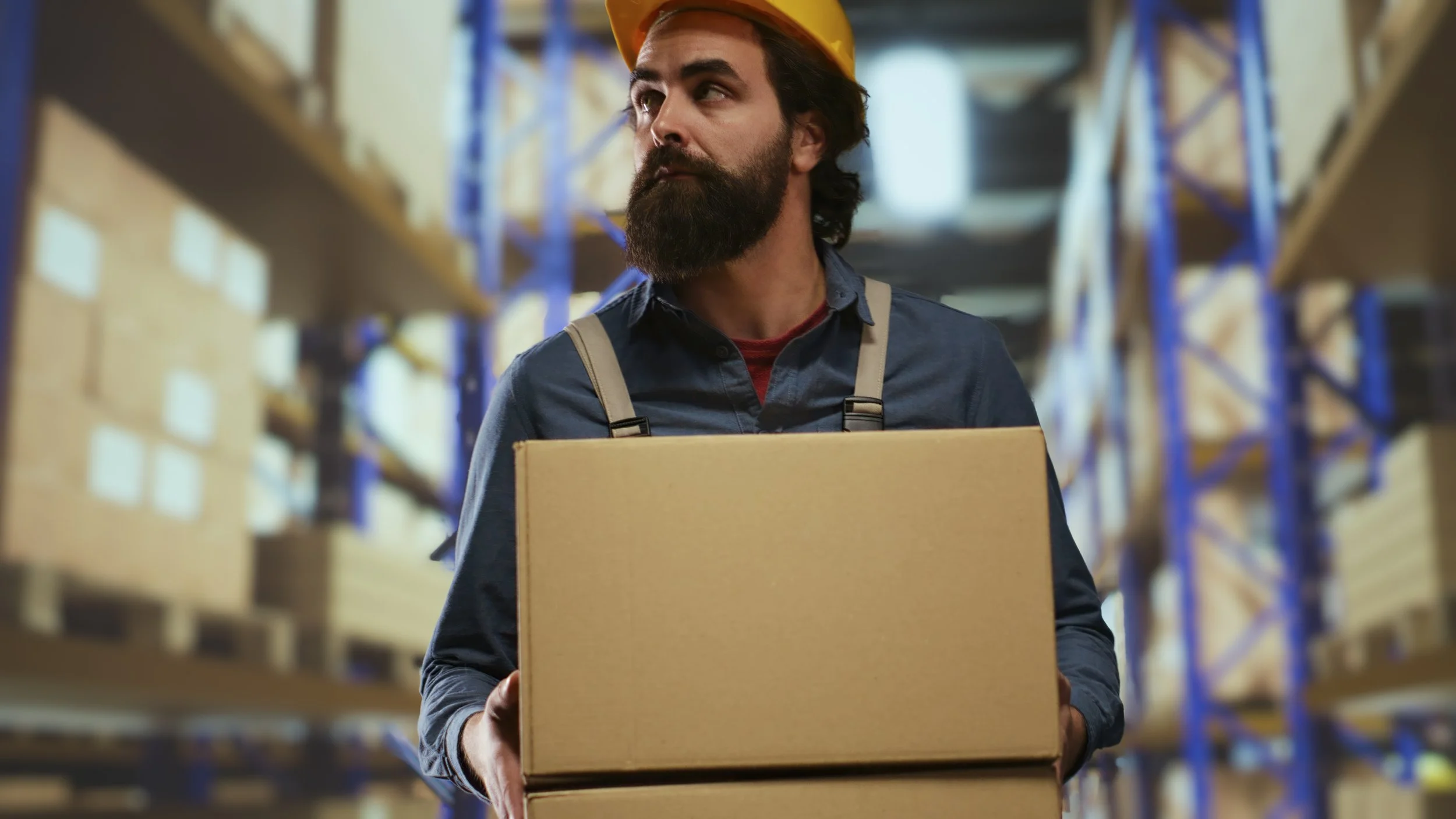 A man with a beard wearing a yellow safety helmet is carrying a cardboard box in a warehouse aisle filled with shelves of neatly stacked boxes.