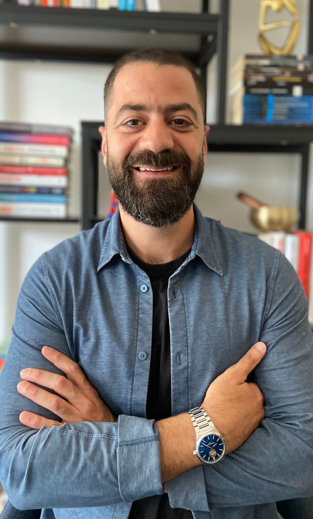 A smiling man with a beard and short hair, wearing a denim shirt and a silver wristwatch, standing in front of a bookshelf with books and decorative items.