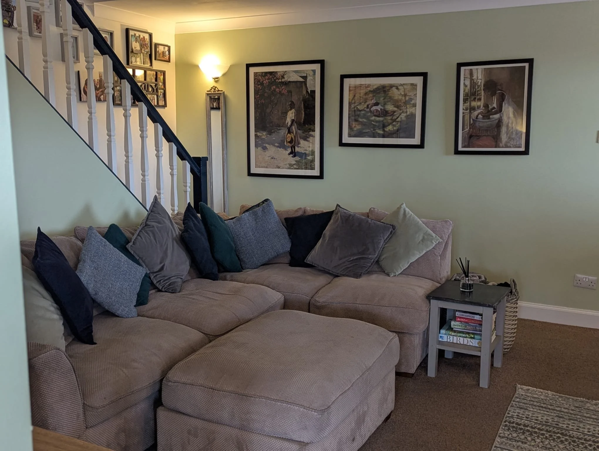 Living room with beige sectional sofa adorned with multiple pillows in various colors and textures, a small black side table with books and a reed diffuser, framed artwork on the wall, and a staircase with a white and black railing.