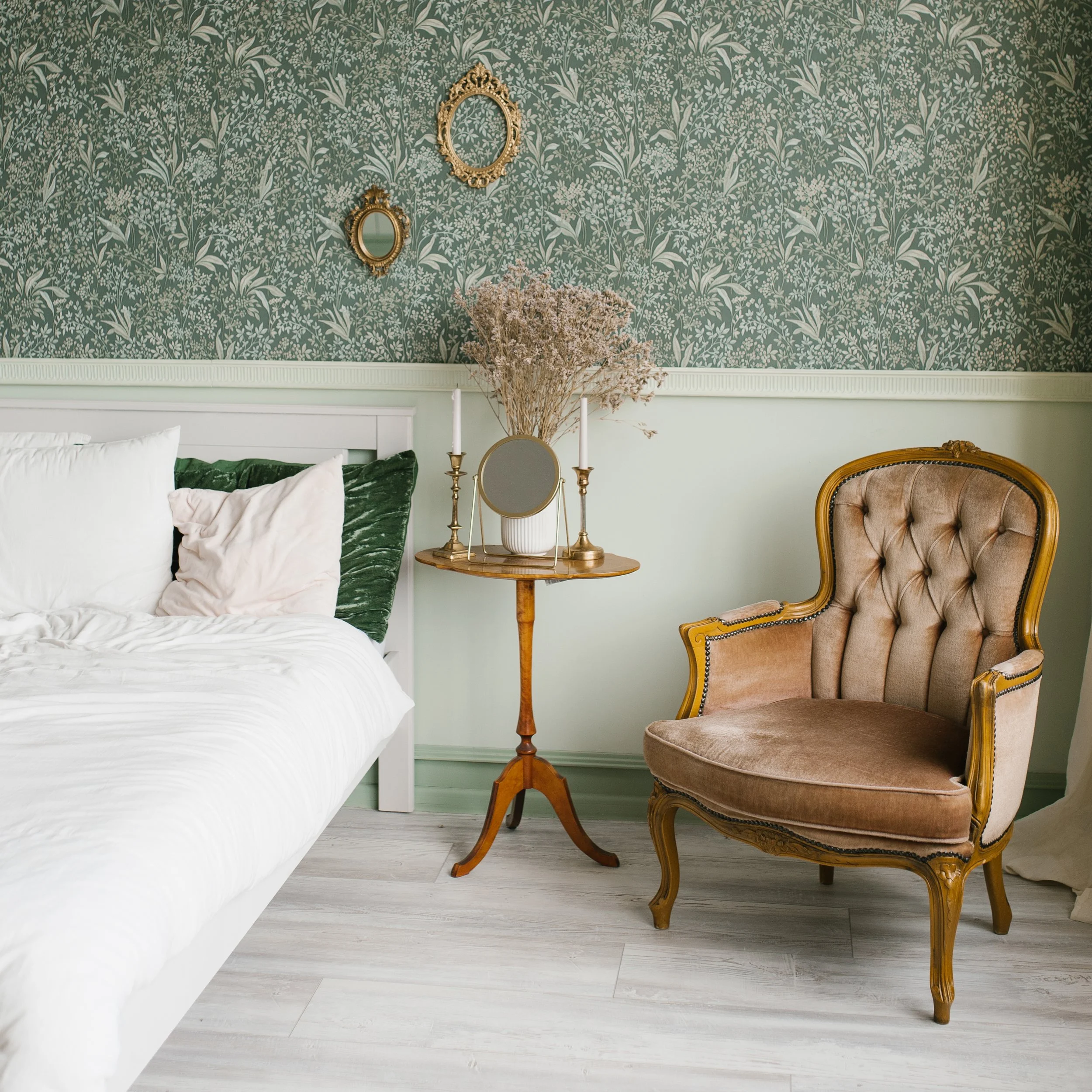 A bedroom with a white bed, green pillows, a vintage armchair with beige upholstery, a wooden side table with a small mirror, dried flowers, and two candlesticks. The background features green floral wallpaper with a white chair rail.