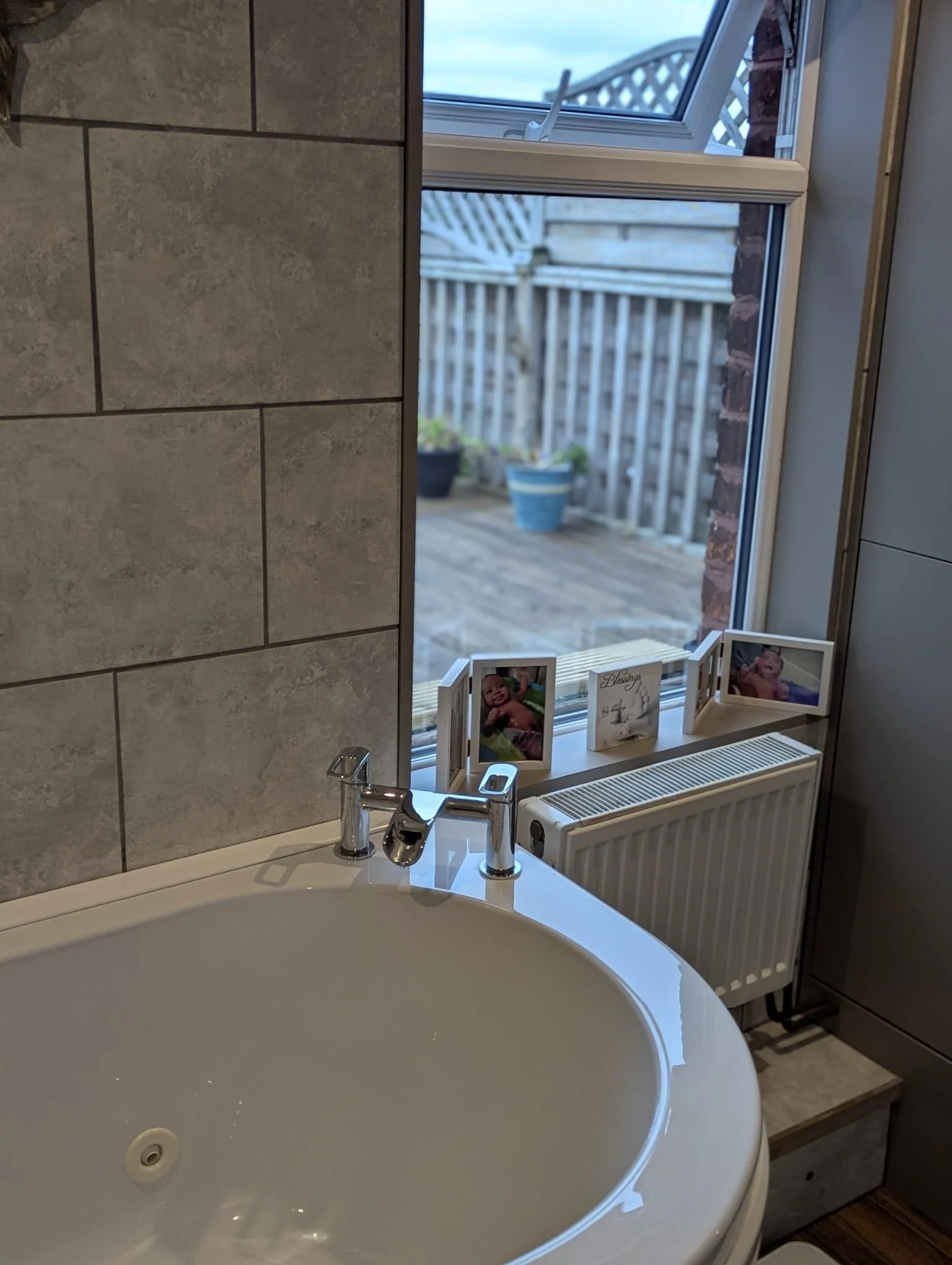A bathroom corner with a white bathtub, a chrome faucet, a window with a view of a backyard patio with potted plants and a wooden fence, and framed photographs on the window sill.