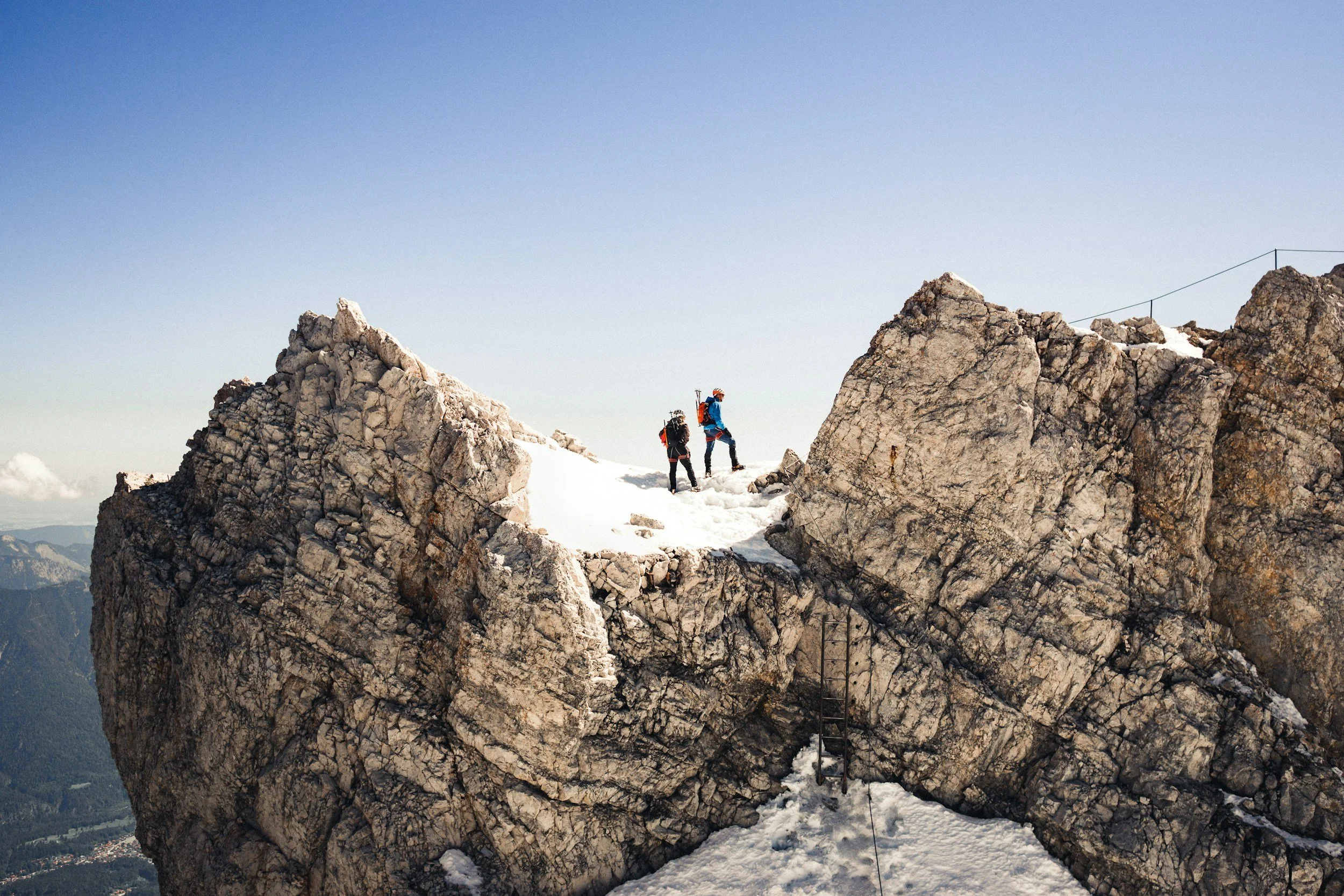 Two hikers in winter gear ascending a snow-covered rocky mountain peak, with a clear sky and distant mountains in the background.
