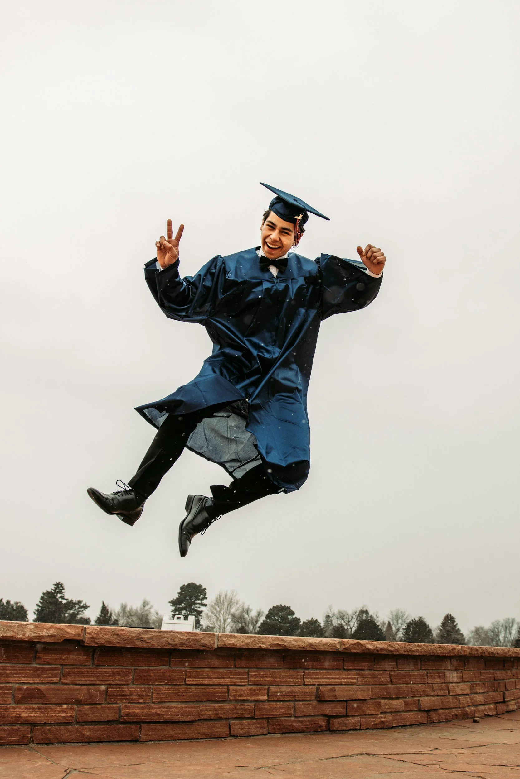 A young man in a graduation cap and gown jumping in the air, smiling, against a cloudy sky, with trees and a brick wall in the background.