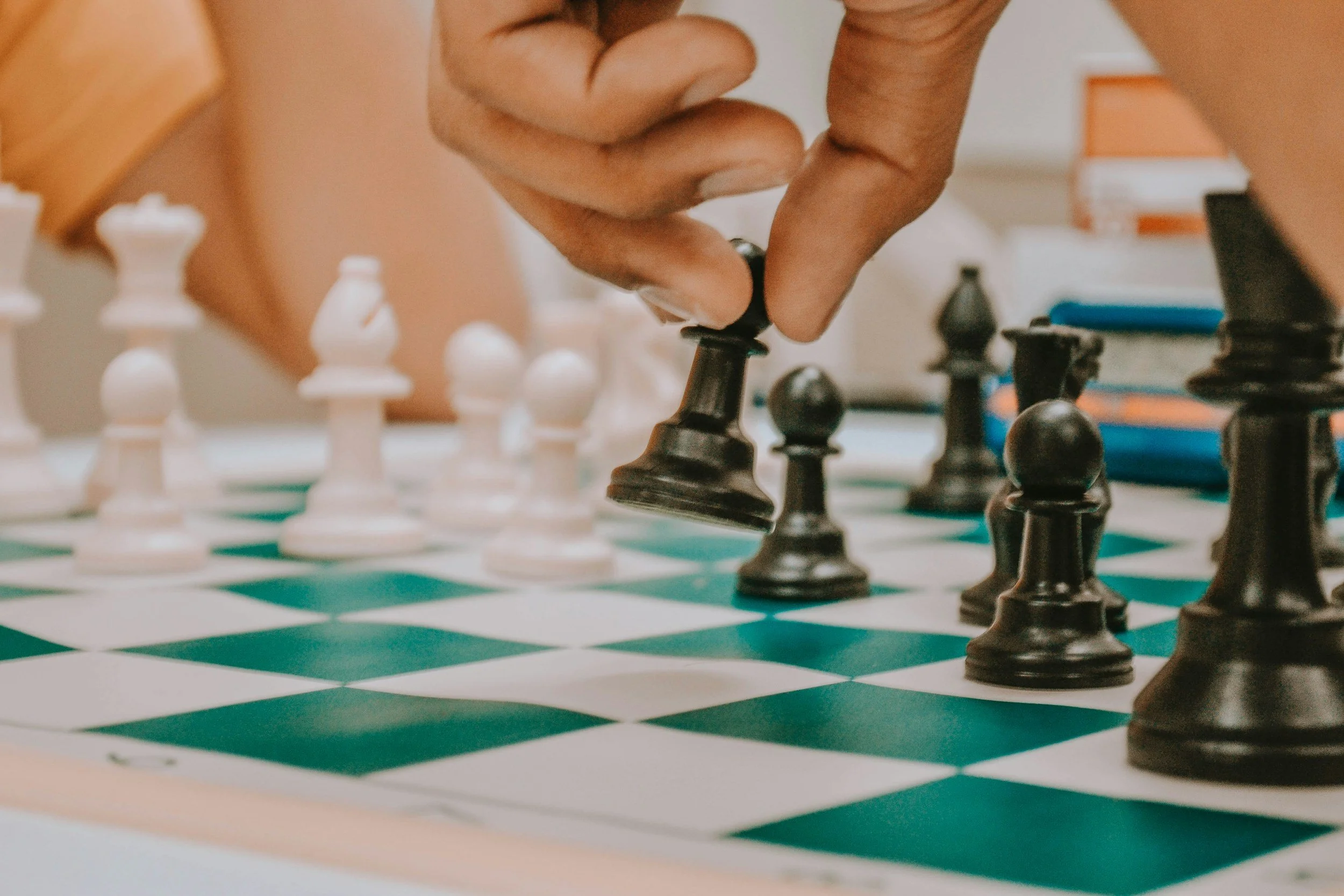 Close-up of a person's hand moving a black chess piece on a green and white chessboard, with white and black chess pieces in the background.