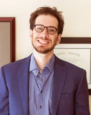 A man with glasses and a beard, wearing a navy blazer and blue checkered shirt, smiling in an indoor setting.