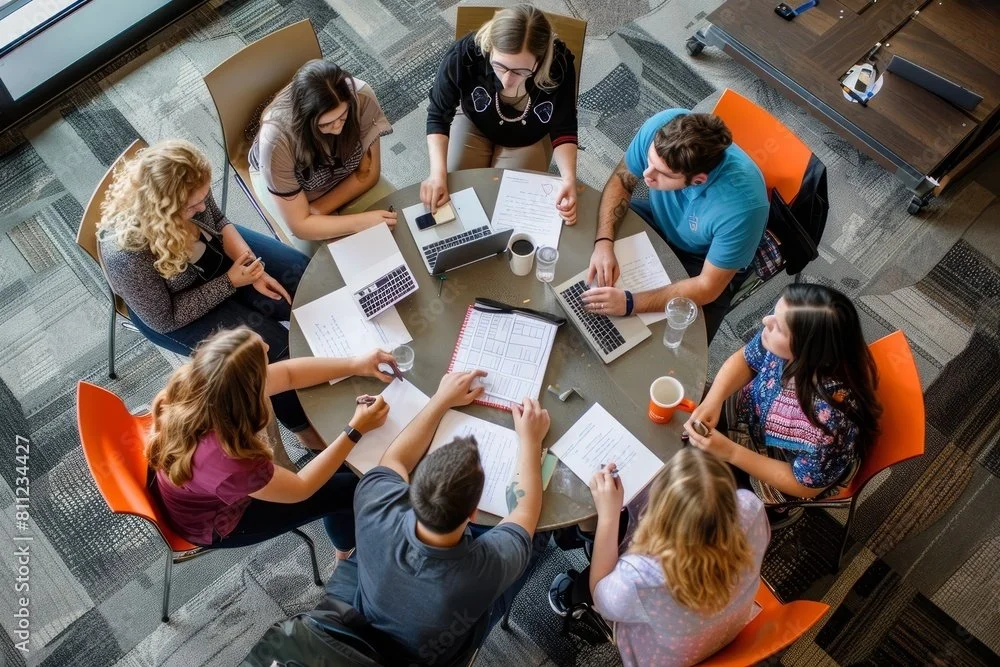 A diverse group of seven people sitting around a round table in a meeting room, working on laptops and reviewing documents.