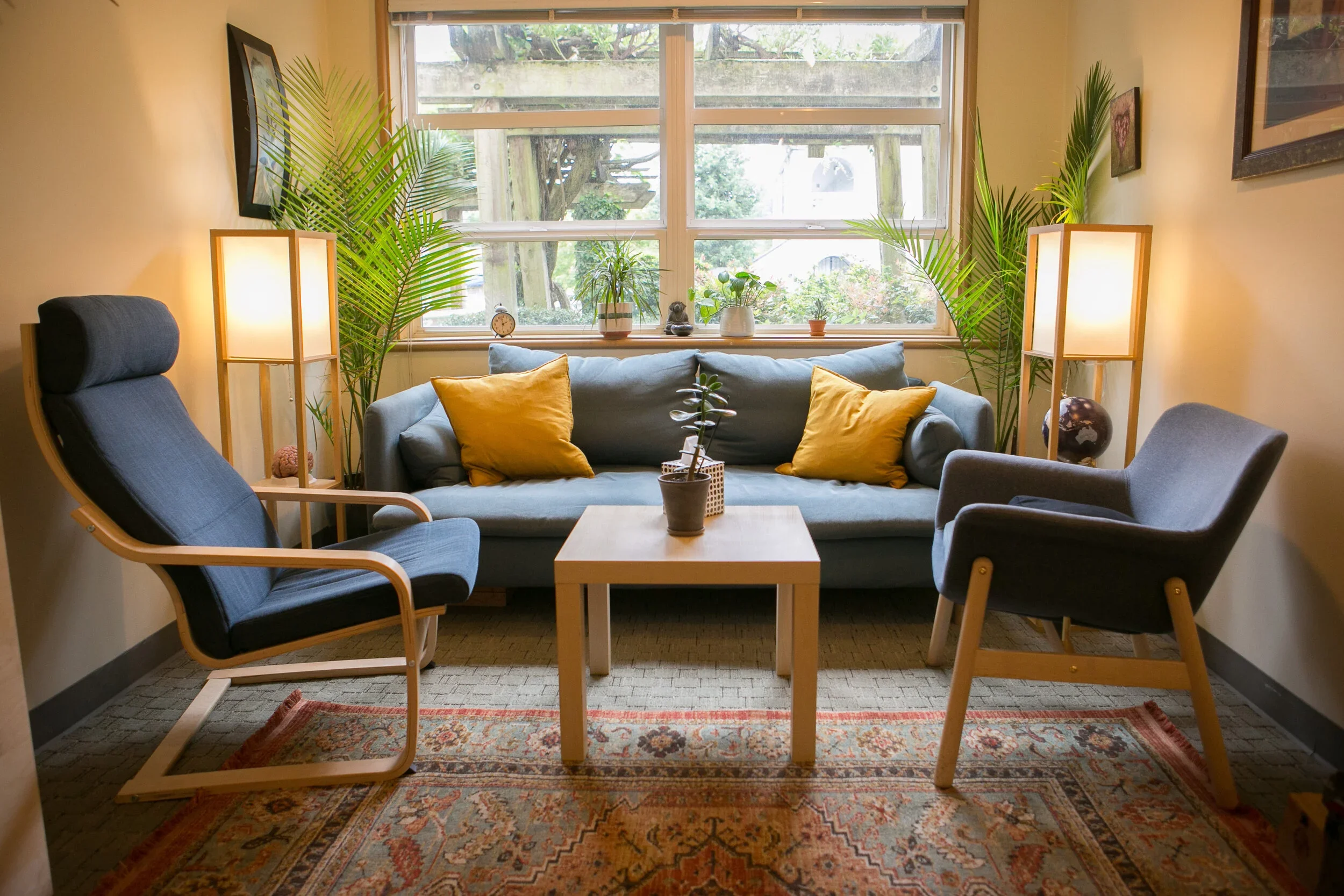 A cozy living room with a large window, a light blue sofa with yellow pillows, two matching armchairs, a wooden coffee table, plants, and lamps on either side. It has a colorful area rug and framed pictures on the walls.
