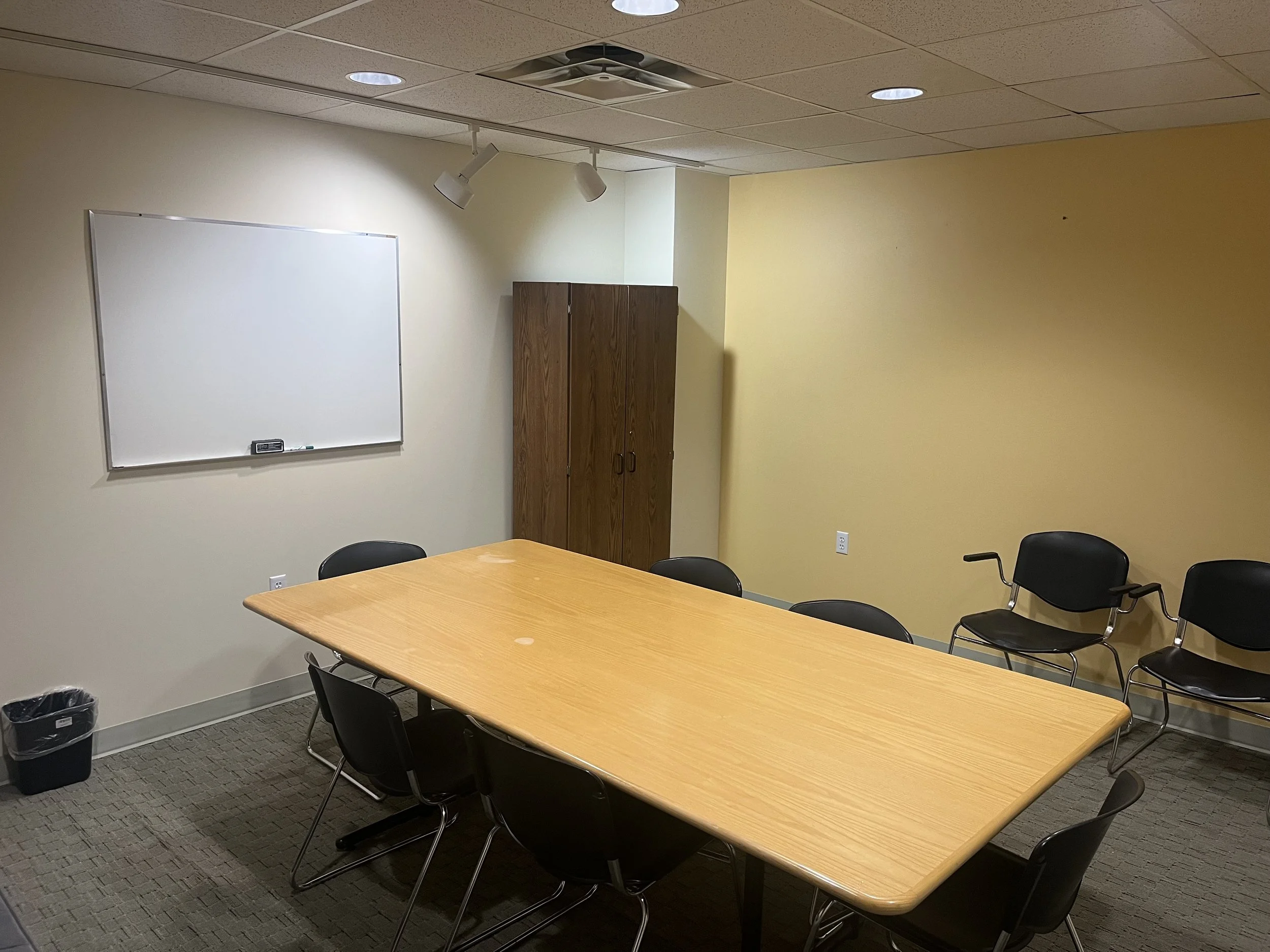 Empty conference or meeting room with a rectangular wooden table, six black chairs around it, a whiteboard on the wall, a brown cabinet in the corner, and a trash can on the floor.