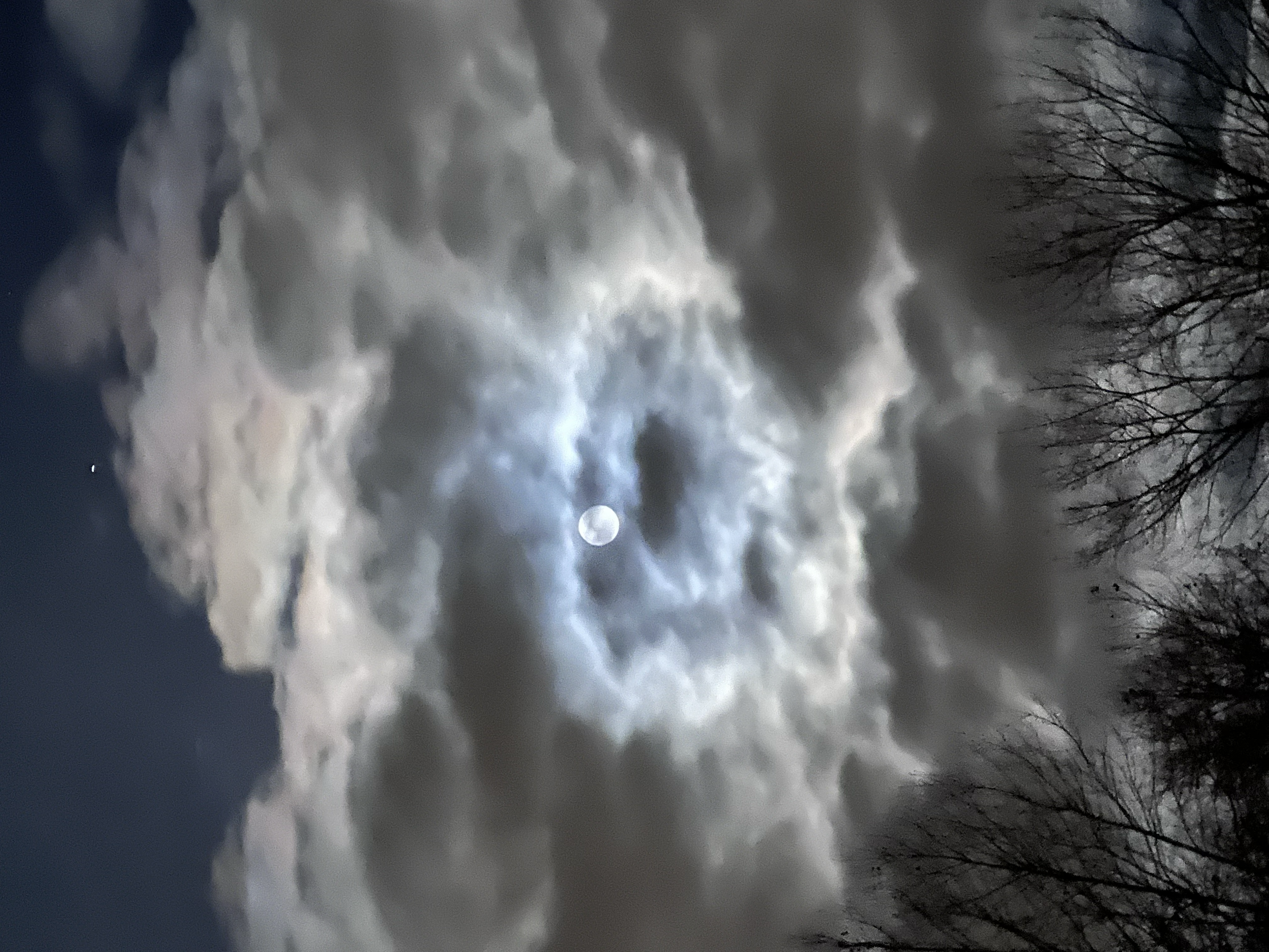 The moon partially obscured by clouds in a night sky, with leafless trees on the right side.