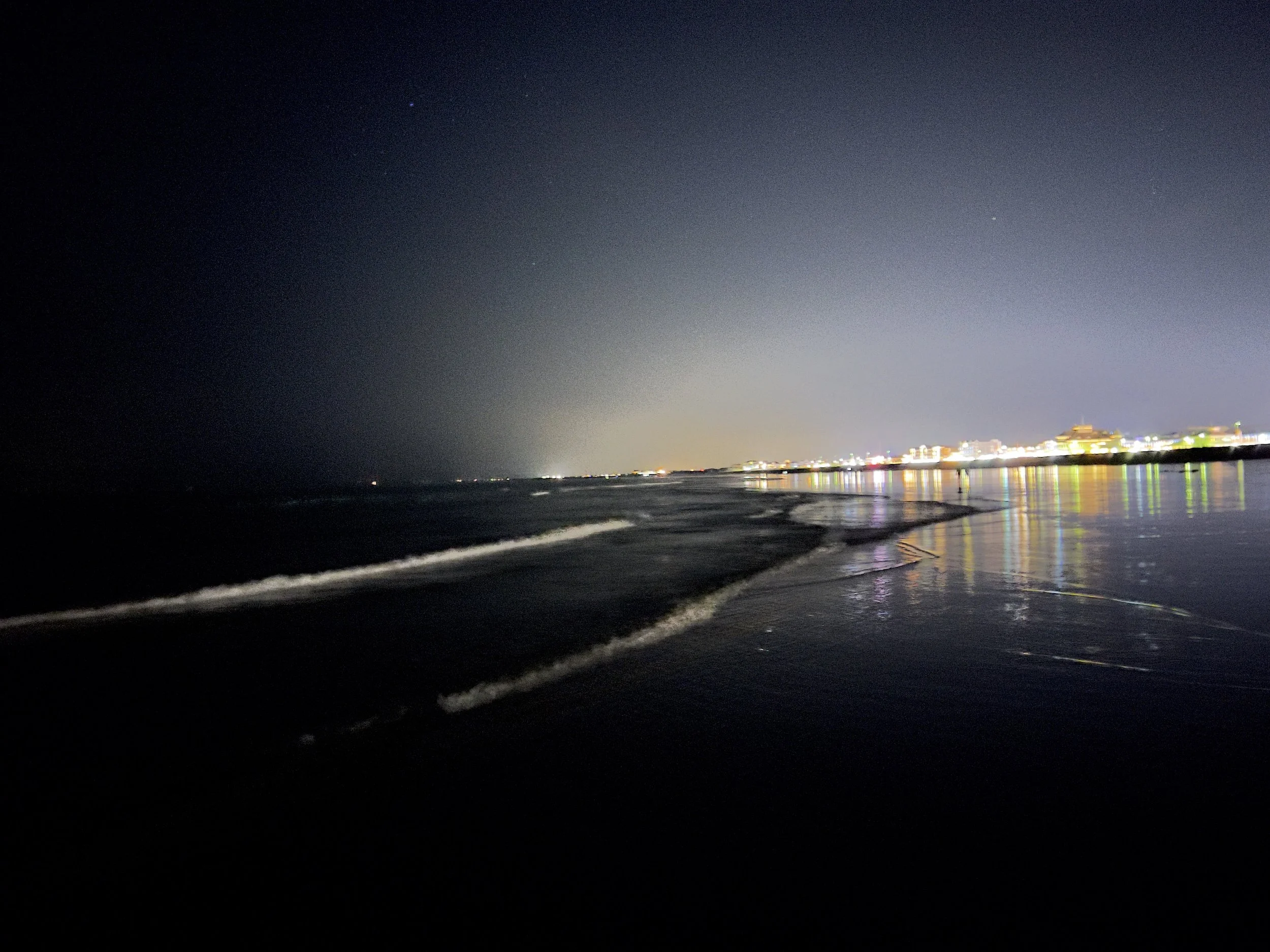 Nighttime view of the beach with gentle waves reflecting city lights in the distance.