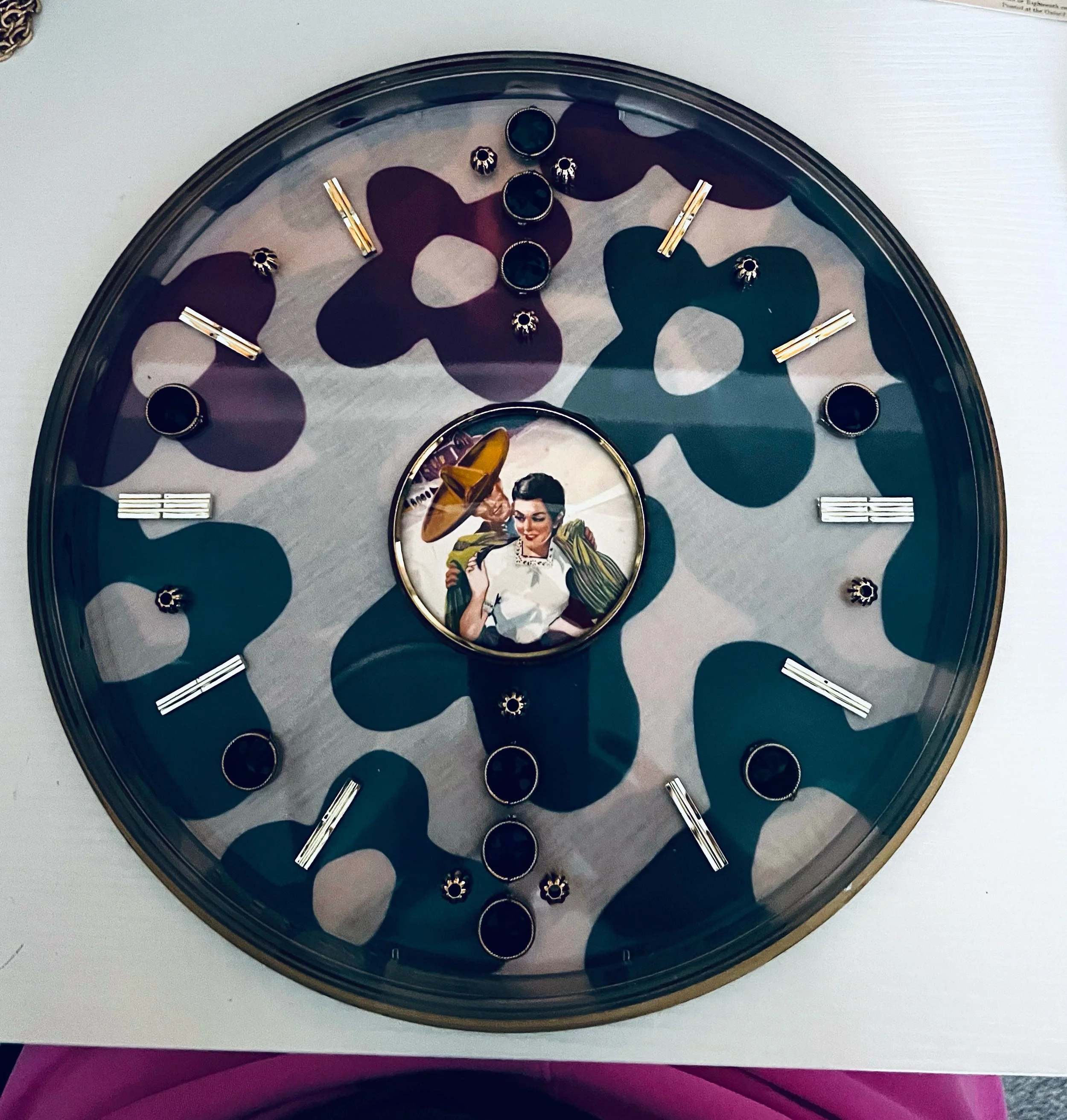A round wall clock with a vintage 1950s style image of a woman in the center, surrounded by a camouflage pattern of dark purple, teal, and beige shapes on the clock face, with black and gold markers at each hour.
