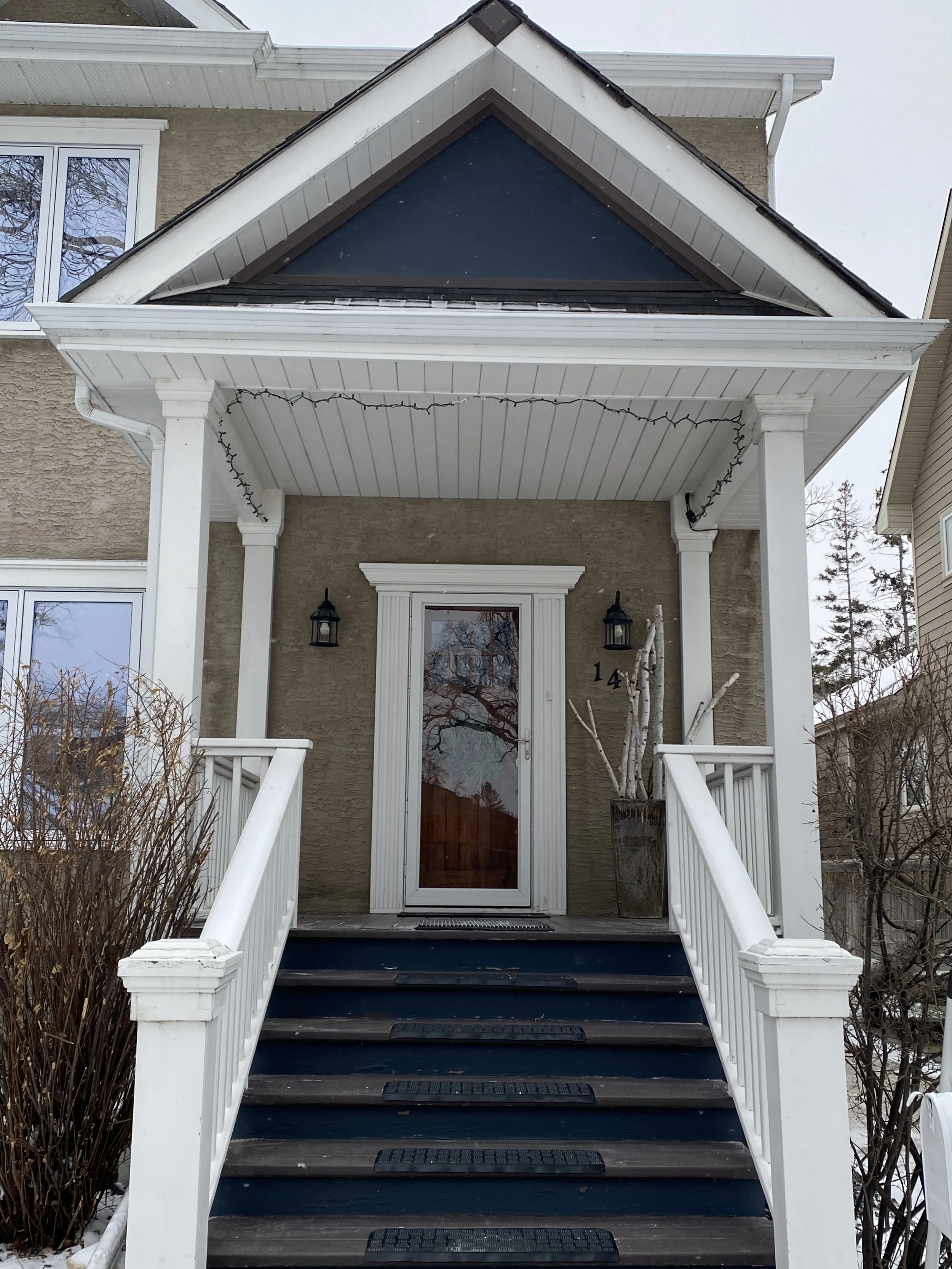 Front porch of a house with dark blue stairs, white railings, and a glass front door, flanked by two black lantern-style lights, with a dried plant in a tall pot on the right and winter bushes on the left, in a snowy setting.