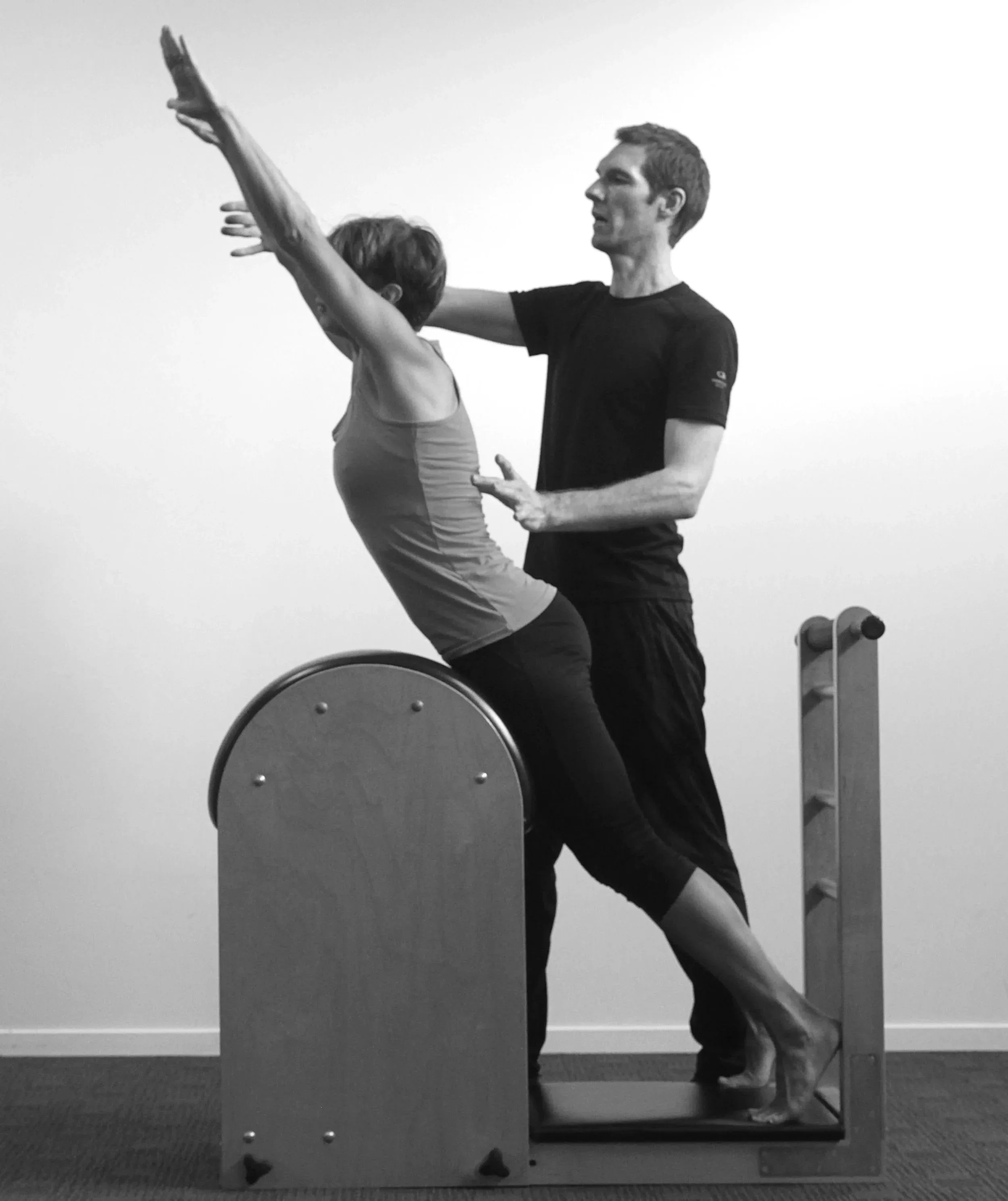 A woman is practicing Pilates with the assistance of Paul Jones on a specialised Pilates ladder barrel, in a studio setting.