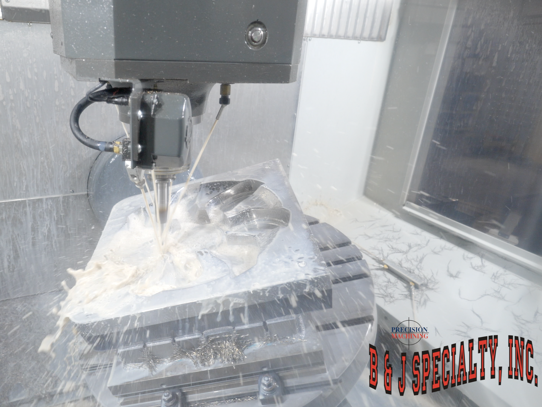 A close-up of a CNC milling machine working on a metal block, cutting out a design with coolant liquid visible. The machine is enclosed in a metallic and transparent case with some text and logos on it.