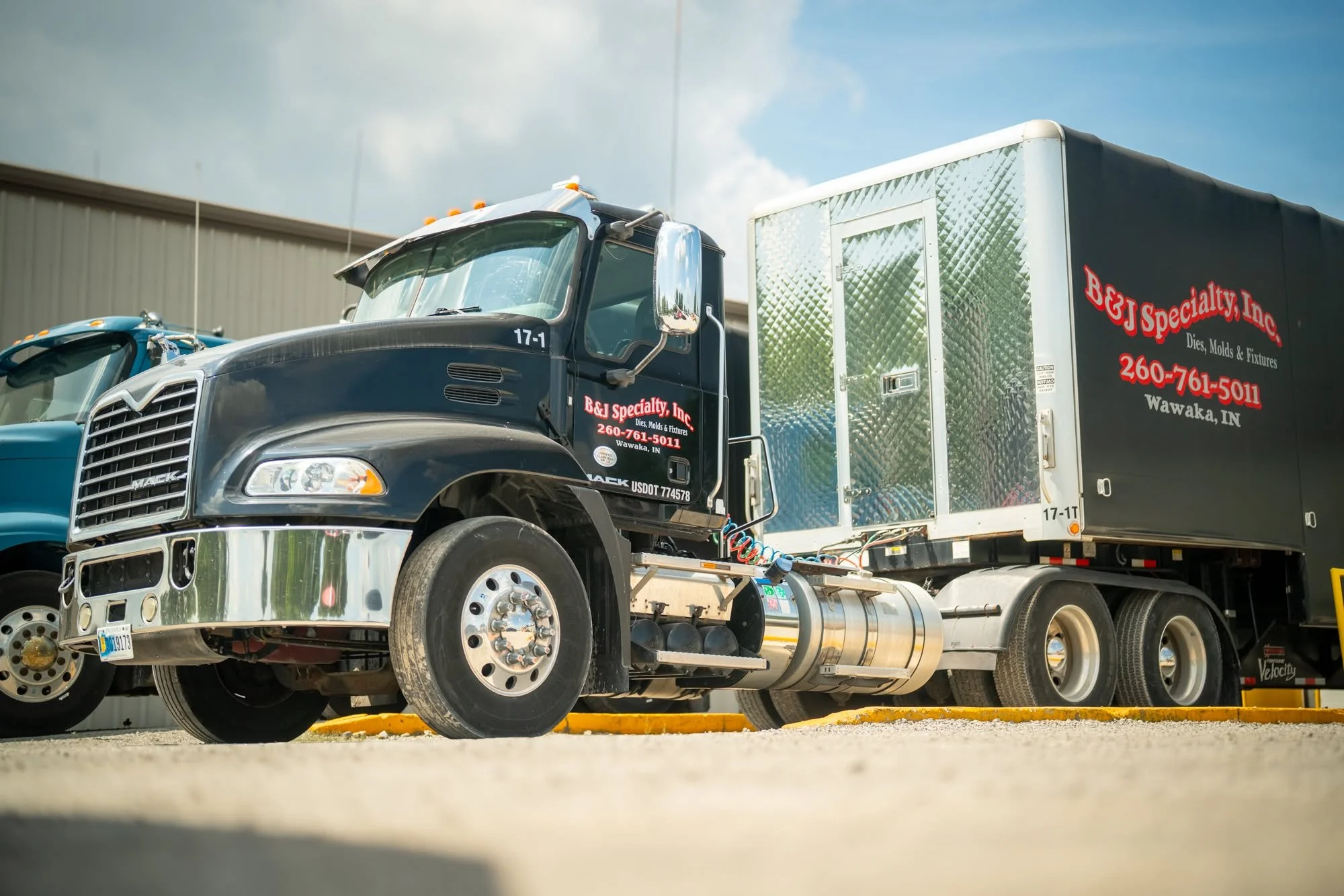 A black Mack semi-truck with a shaded logo on the door, hauling a large black trailer with red and white lettering advertising B&J Specialty, Inc., a company in Wawaka, Indiana, that deals with dies, molds, and fixtures. The truck is parked on a concrete surface with a warehouse building in the background and cloudy skies above.