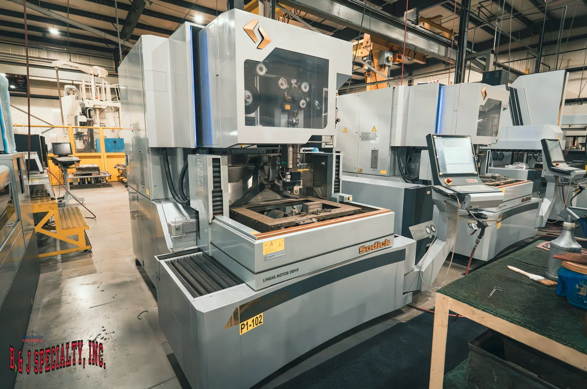 Industrial manufacturing factory with large Sodick CNC machine, workbenches, computer stations, and industrial equipment in a spacious warehouse.