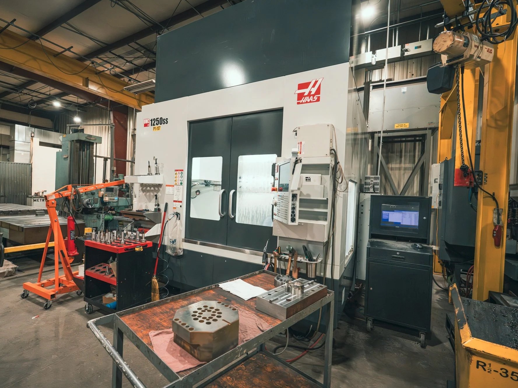 Industrial workshop with a large CNC machine, work tables, tools, and equipment for manufacturing or machining processes.