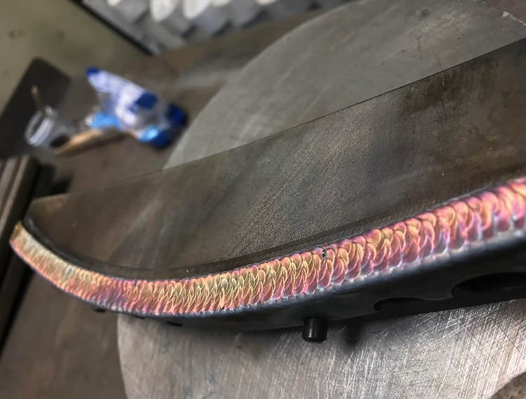 Close-up of a metal workpiece with a colorful, heat-affected zone from welding or grinding, resting on a workbench with workshop tools and materials in the background.