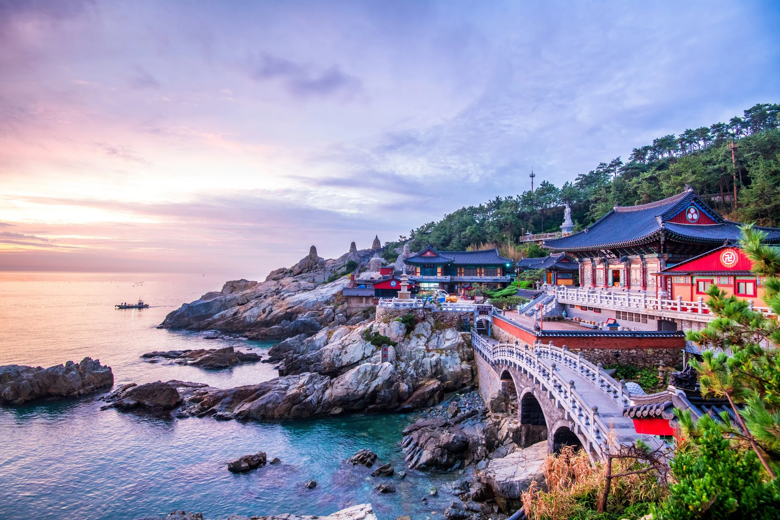Scenic view of a traditional Asian temple complex on a rocky coastline at sunset, with pink and purple hues in the sky, a boat on the water, and lush green trees on the hillside.