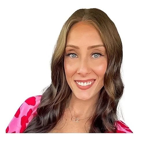 Portrait of a woman with long brown hair, blue eyes, and a bright smile, wearing a pink floral top and a gold necklace, against a white background.
