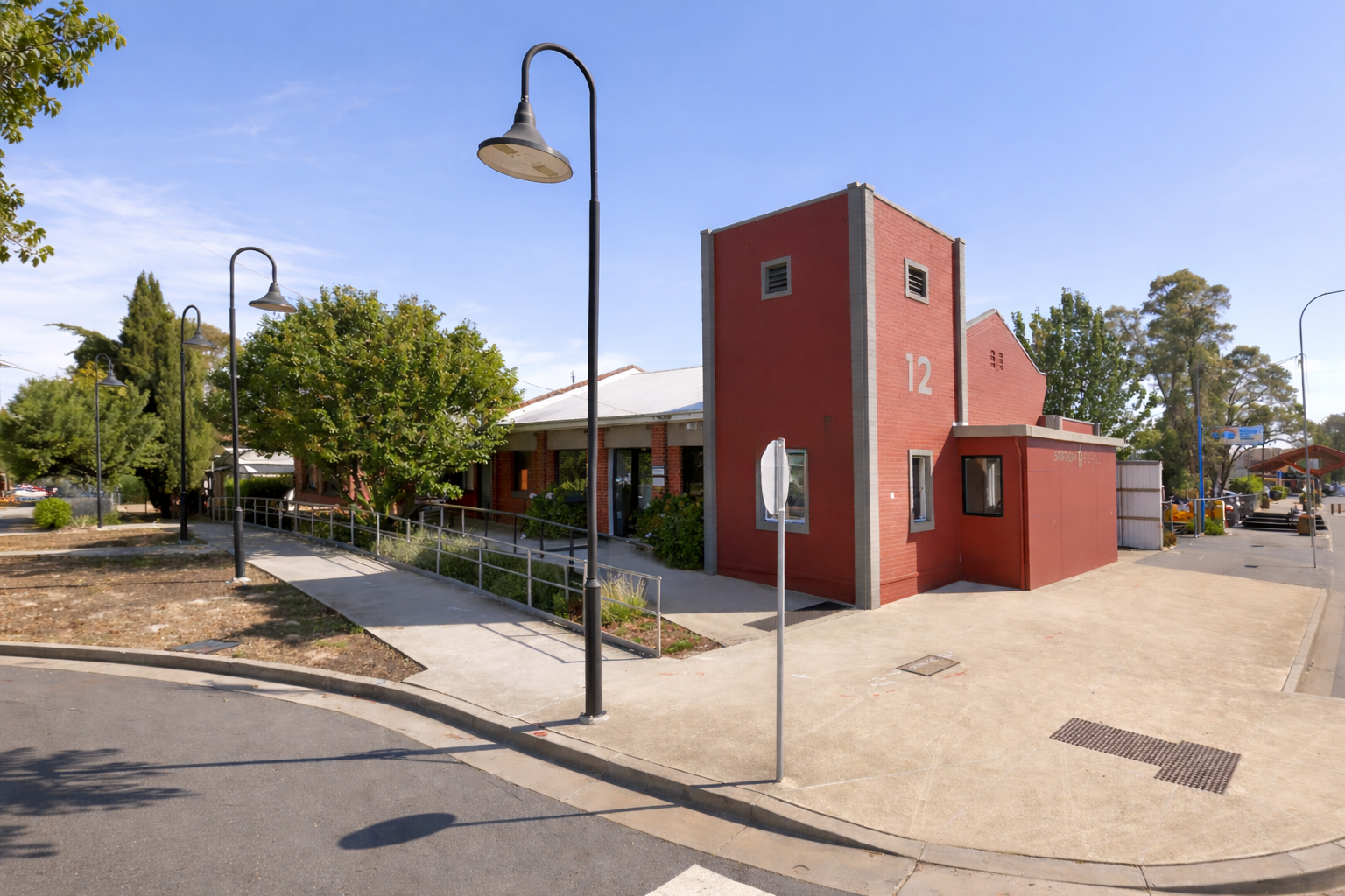 A red brick building with a tall rectangular tower marked with the number 12, situated on a sidewalk with street lamps and trees nearby.