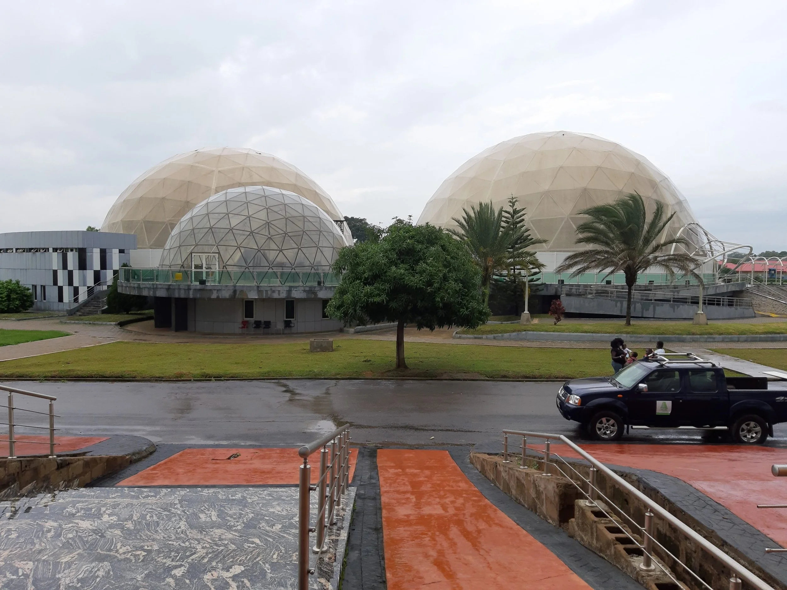 A group of geodesic domes with palm trees in front, a grassy area, a wet road, and a black pickup truck parked nearby.