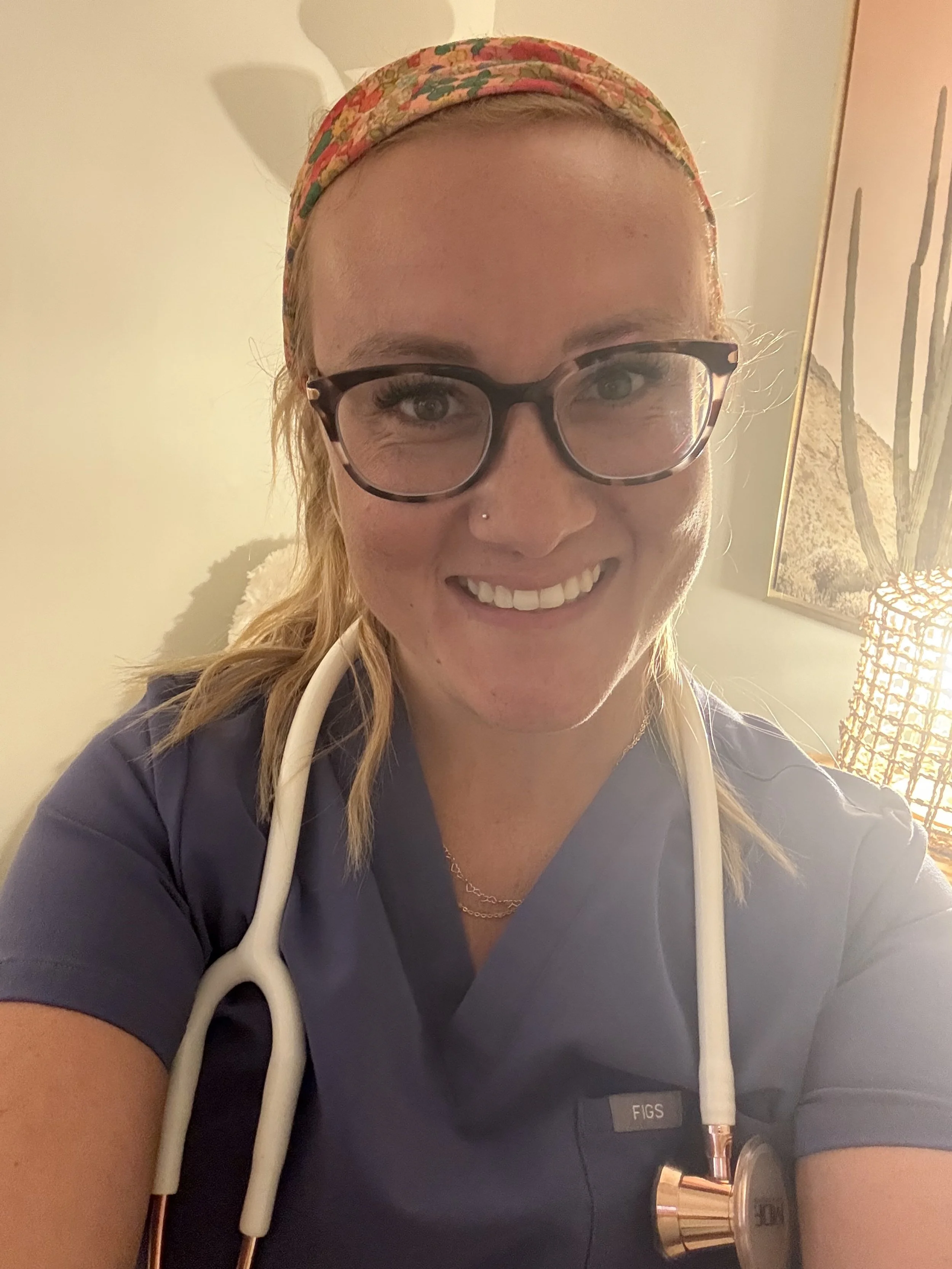 A woman with blonde hair, wearing glasses, a colorful headband, and blue scrubs with a stethoscope around her neck, smiling at the camera.